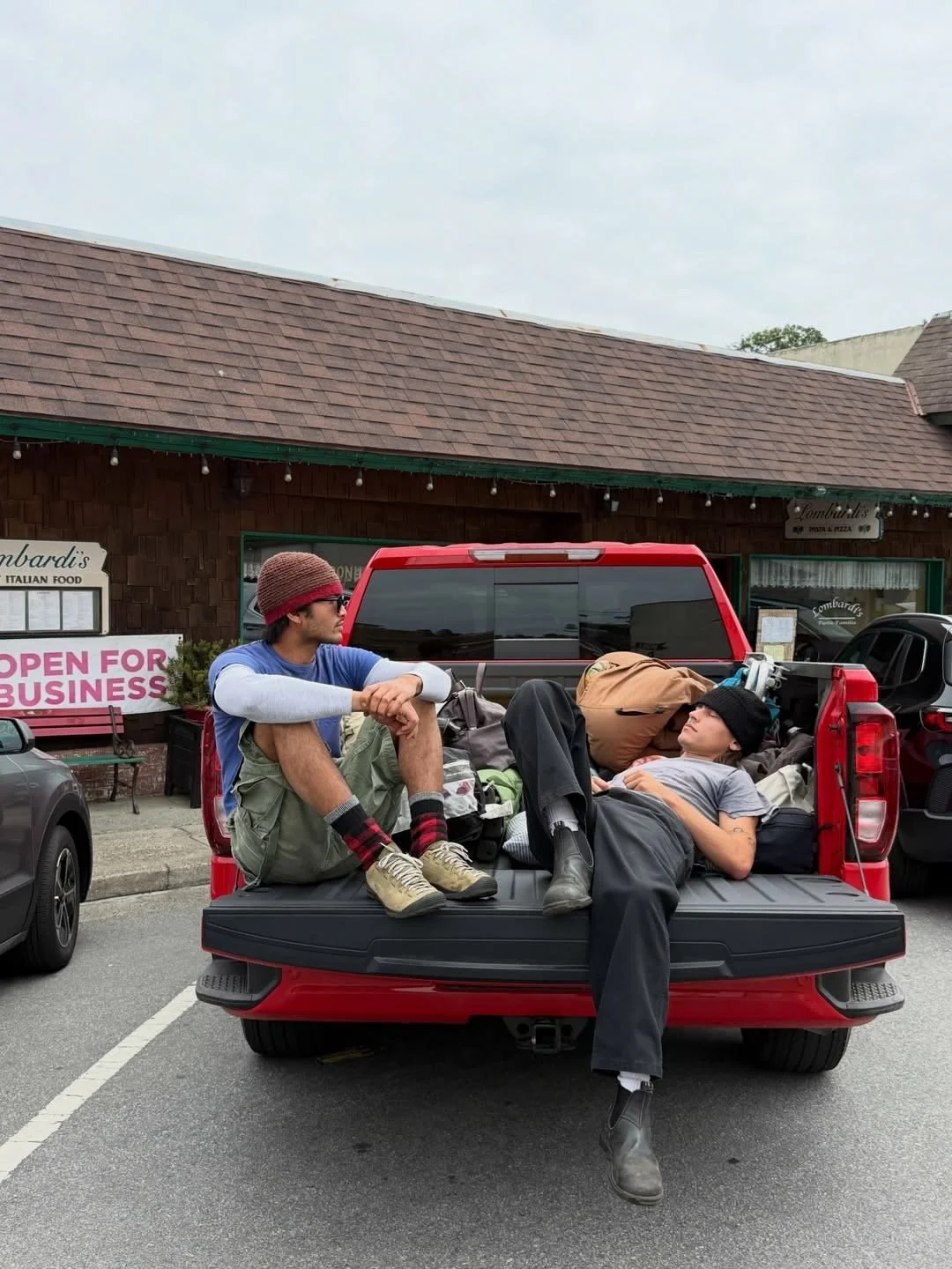 Two young people resting in the bed of a red pickup truck in a parking lot, surrounded by luggage, in front of a restaurant with a sign that reads 'OPEN FOR BUSINESS'.