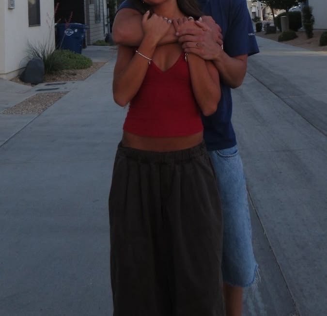 A woman wearing a red sleeveless top and dark pants being embraced from behind by a person in a blue shirt, standing on a sidewalk in a residential neighborhood.
