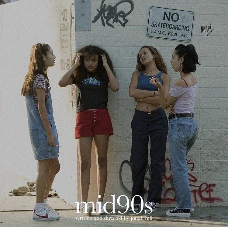 Four young women stand on a sidewalk next to a white brick wall with graffitti and a sign that says 'No Skateboarding'. The women are casually dressed, and they seem to be talking to each other. The photo has a vintage 90s aesthetic.