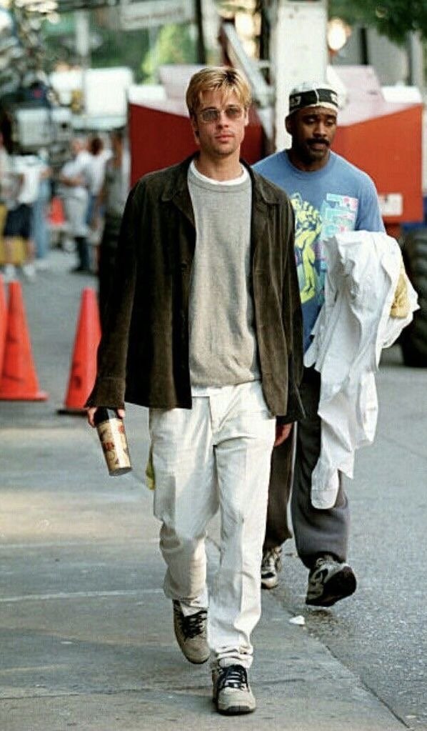 Two young men walking on a city sidewalk, one in a brown jacket and light-colored pants holding a drink, the other in a blue T-shirt with a headband and holding a white jacket.