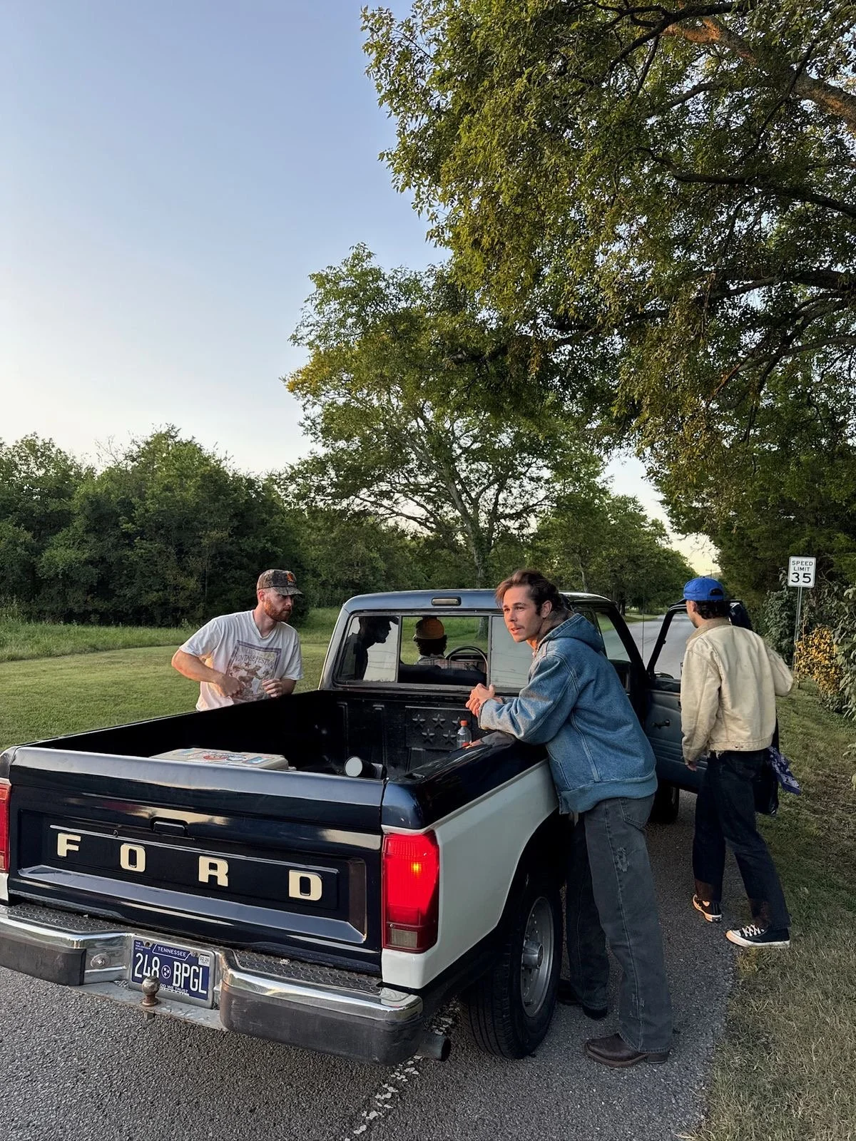 Four young men are gathered around a black and white Ford pickup truck parked on the side of a rural road. Three of them are outside the truck, and one is inside the driver's seat. They are talking and possibly preparing to drive or load items. The s