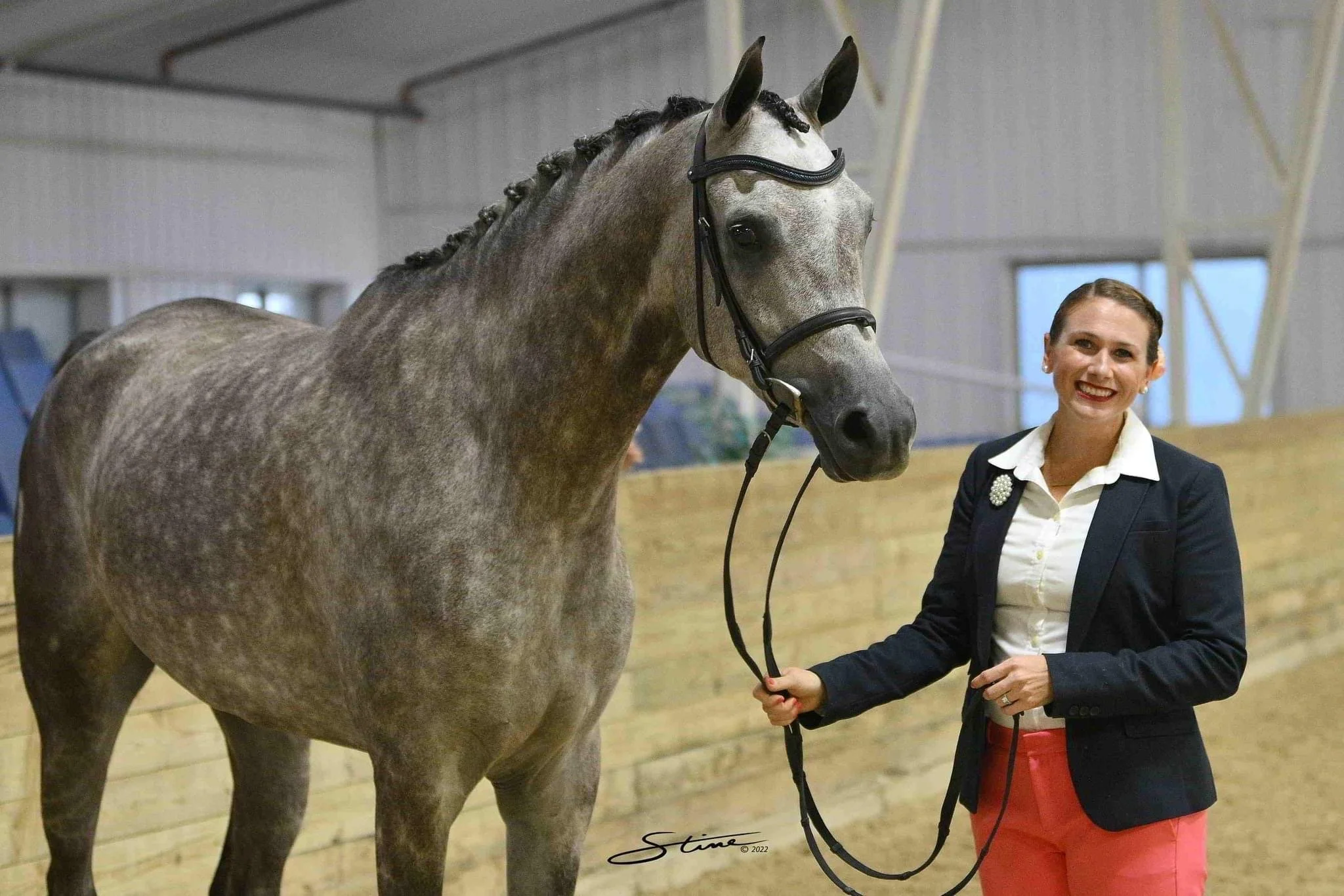 A woman in a black blazer, white shirt, and red pants standing inside an indoor riding arena next to a gray horse with a braided mane. The woman is smiling and holding the horse's reins while the horse looks directly at the camera. The background shows wooden walls and windows.