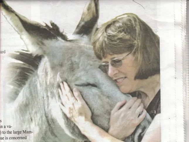 A woman smiling and holding a donkey's face close to her face, showing affection.