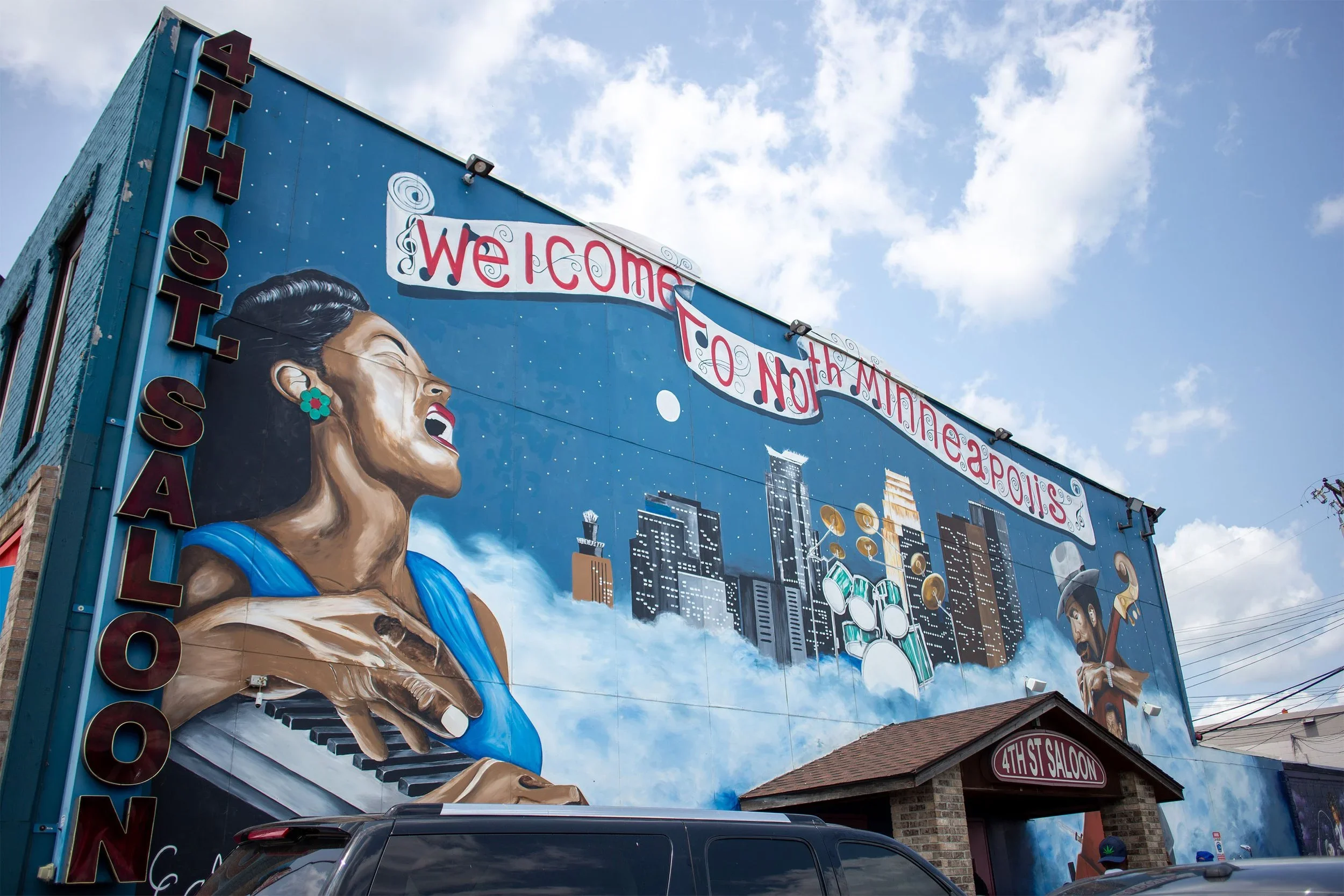 Large mural on the side of a building welcoming visitors with an animated woman playing piano, cityscape, and musical instruments, with a 'WELCOME' sign, under a partly cloudy sky.