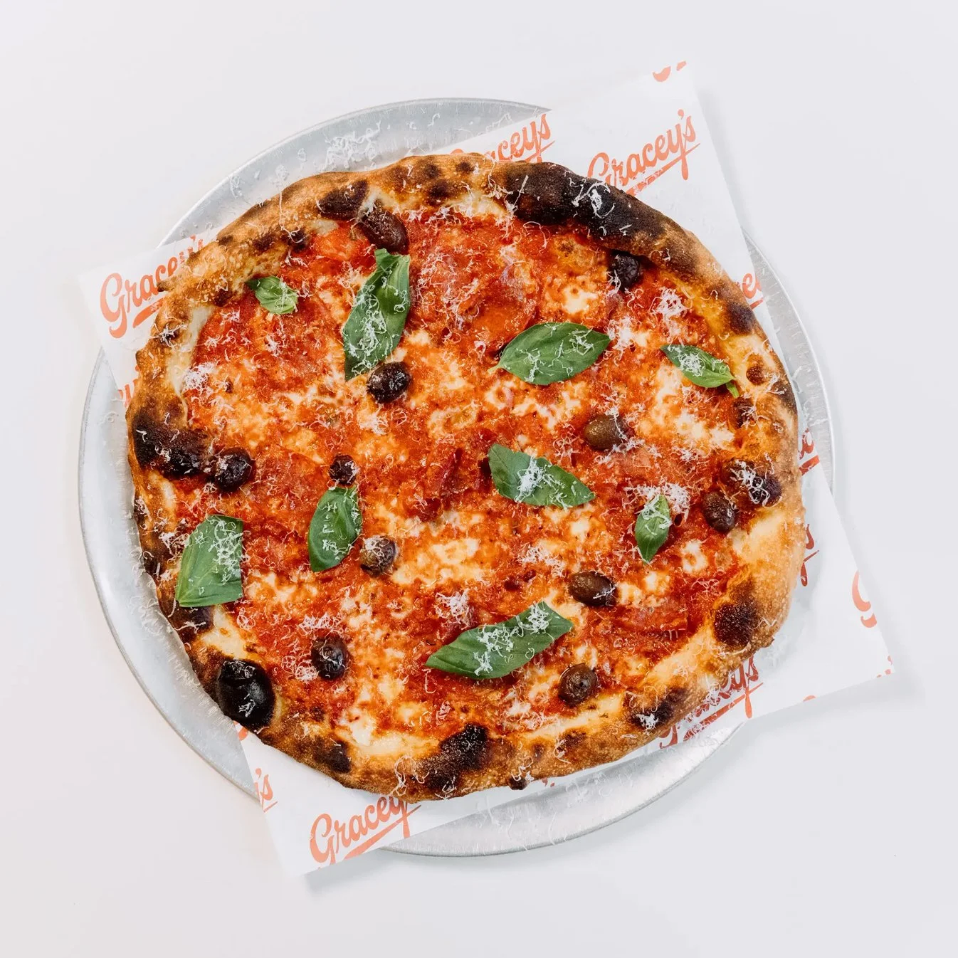 A pizza with tomato sauce, black olives, and fresh basil leaves on top, served on a metal tray lined with branded paper.
