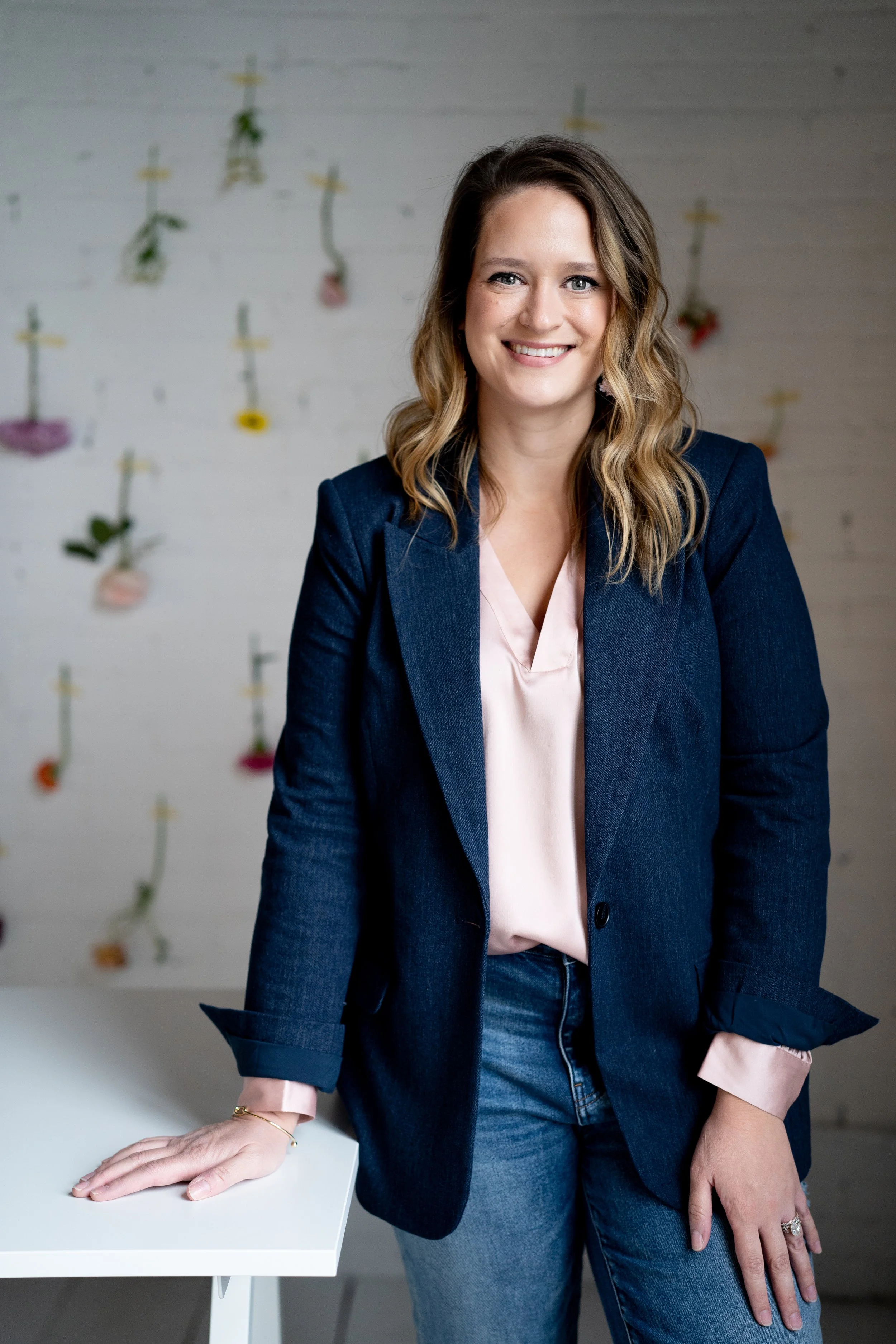 A woman with long wavy hair wearing a navy blazer over a light pink blouse, standing next to a white table with one hand resting on it, in a room with a white brick wall decorated with small hanging flower arrangements.
