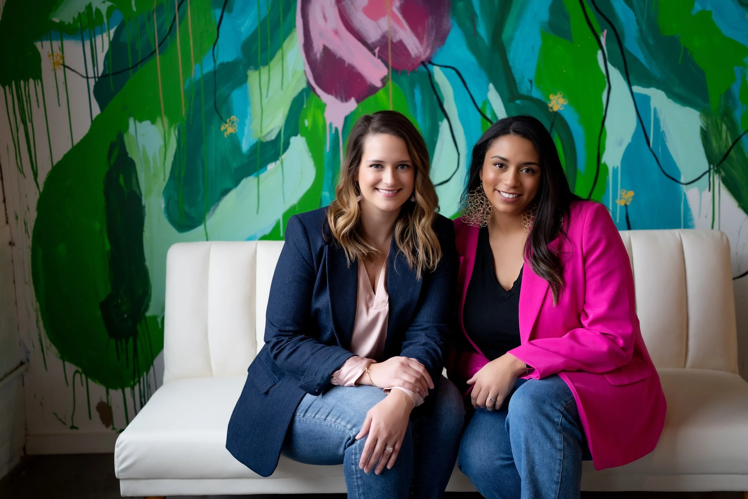 Two women sitting on a white sofa in front of a colorful abstract mural. One woman is wearing a dark blazer over a light pink blouse, and the other woman is wearing a bright pink blazer over a black top and jeans. They are smiling and sitting close together.