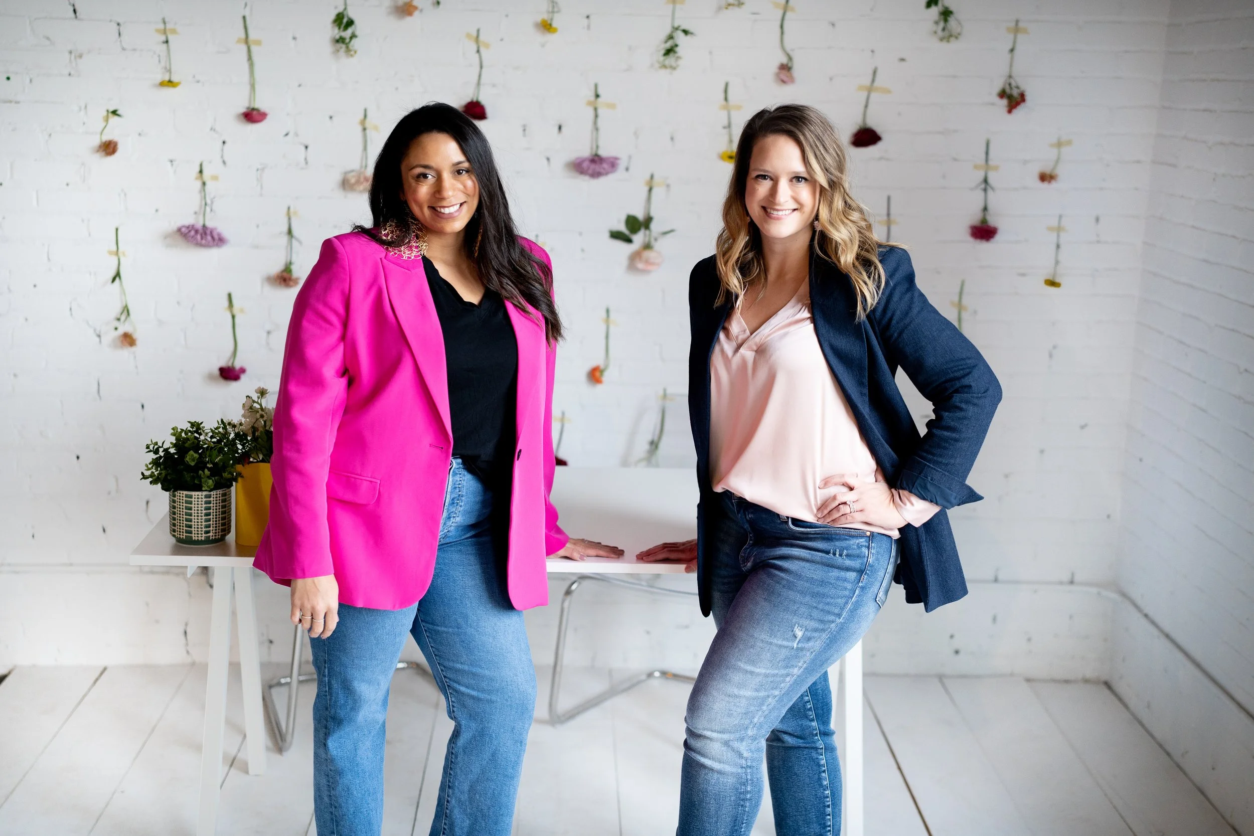 Two women standing in a bright room with a white brick wall decorated with hanging flowers. One woman is wearing a pink blazer and jeans, the other a navy blazer and jeans. They are smiling and posing for the photo.