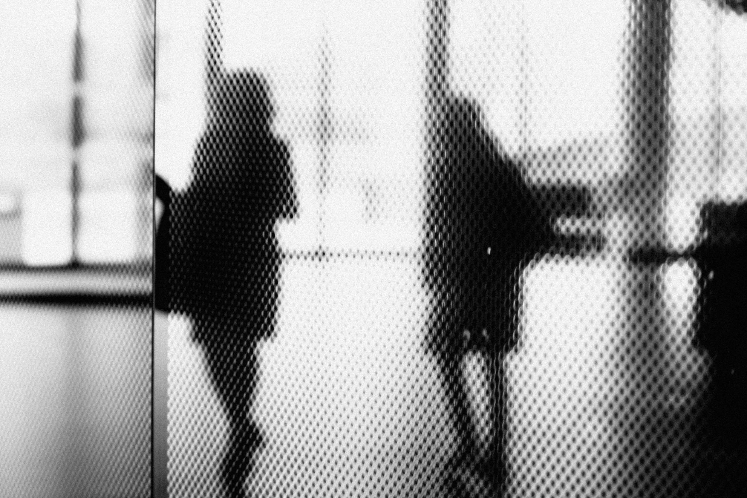 Black and white photo of two people walking in an indoor space, seen through a perforated glass or screen.