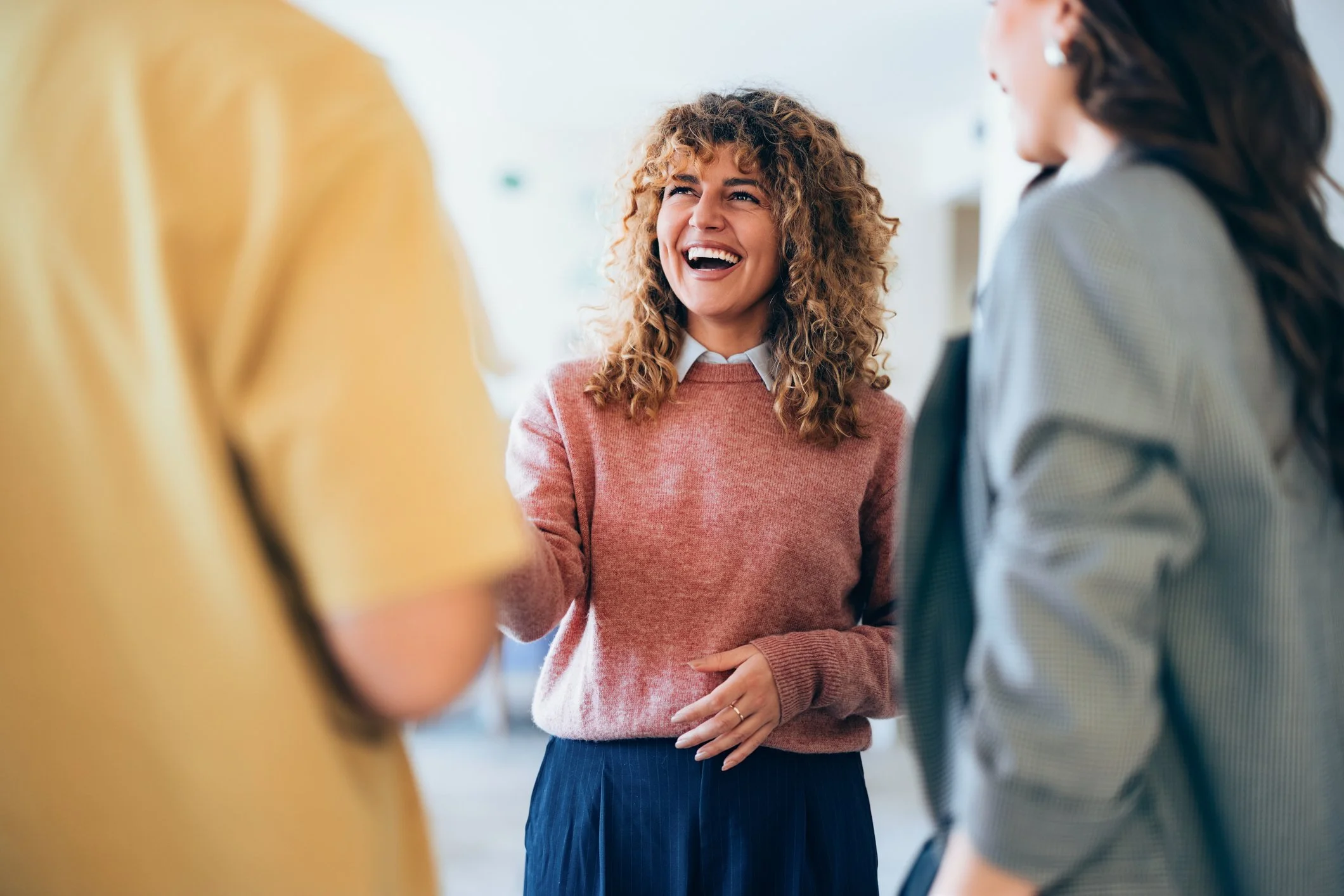 A woman with curly hair and a pink sweater laughing and engaging in conversation with friends in a social setting.