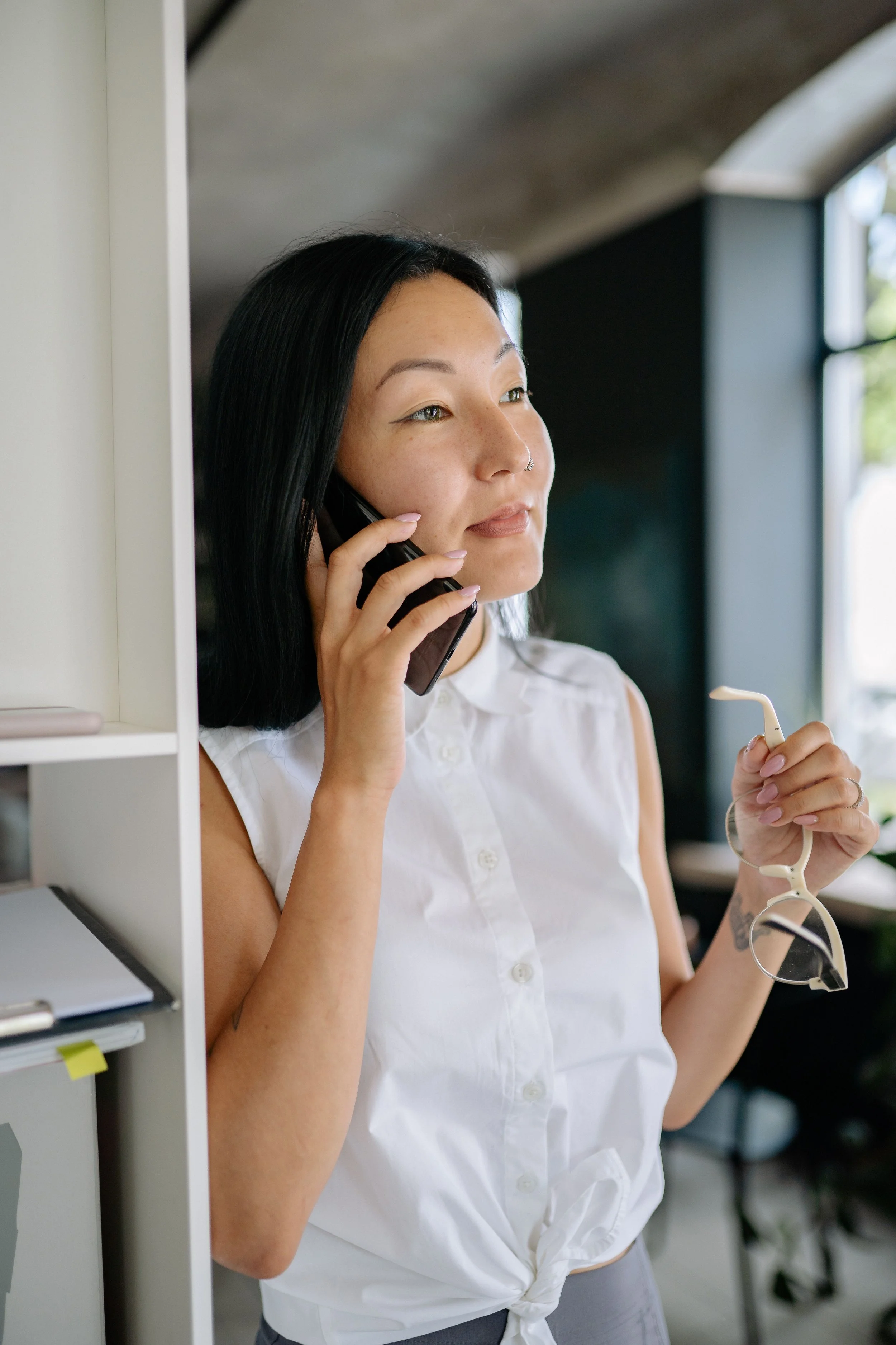 Woman with black hair and a nose ring talking on a cellphone inside a modern office, holding glasses in her left hand, wearing a white sleeveless shirt knotted at the waist.