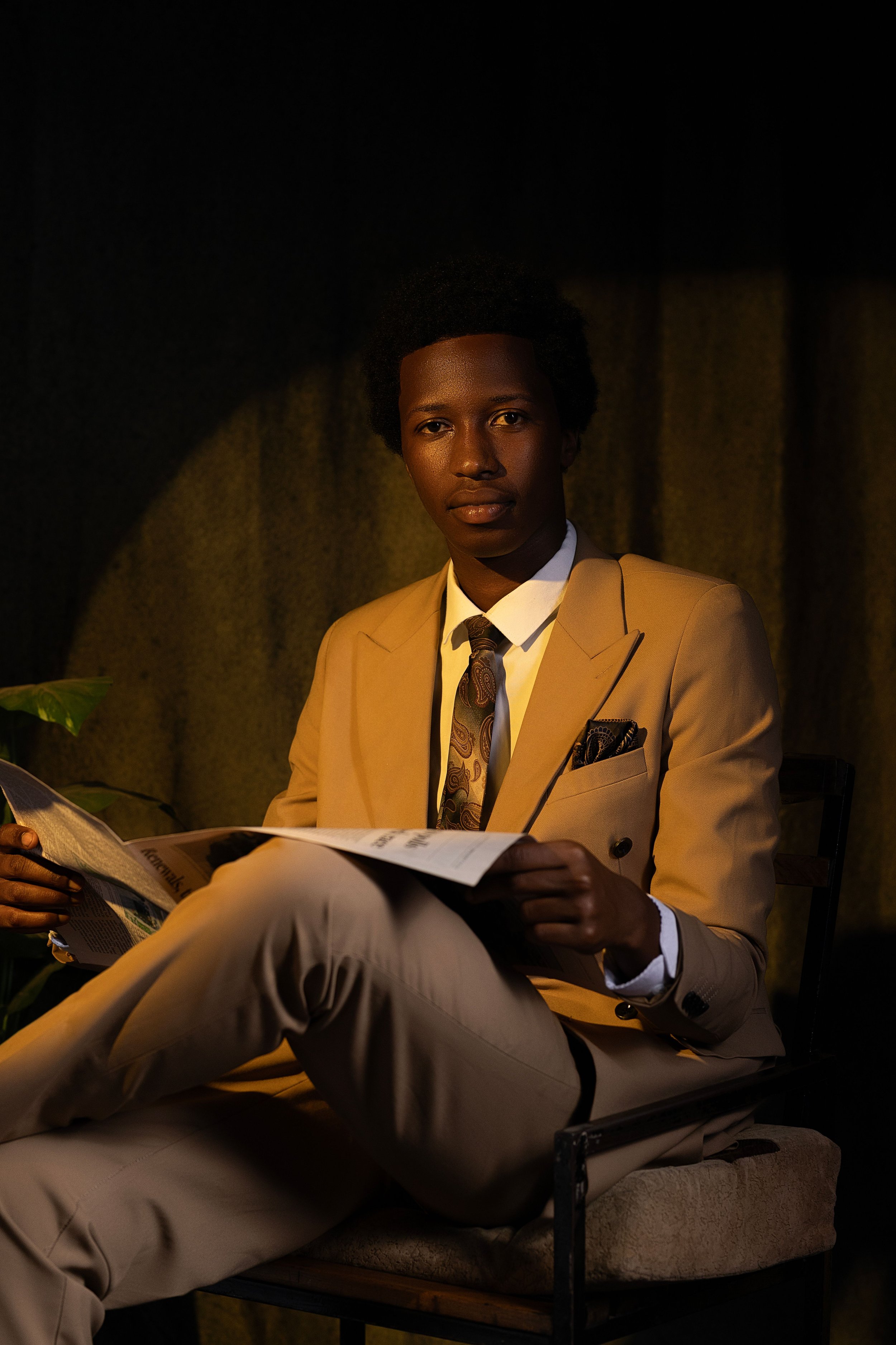 A man with dark skin and short curly hair, wearing a beige suit, white shirt, and patterned tie, sitting in a dimly lit room, reading a newspaper, with a potted plant partially visible in the lower left corner.