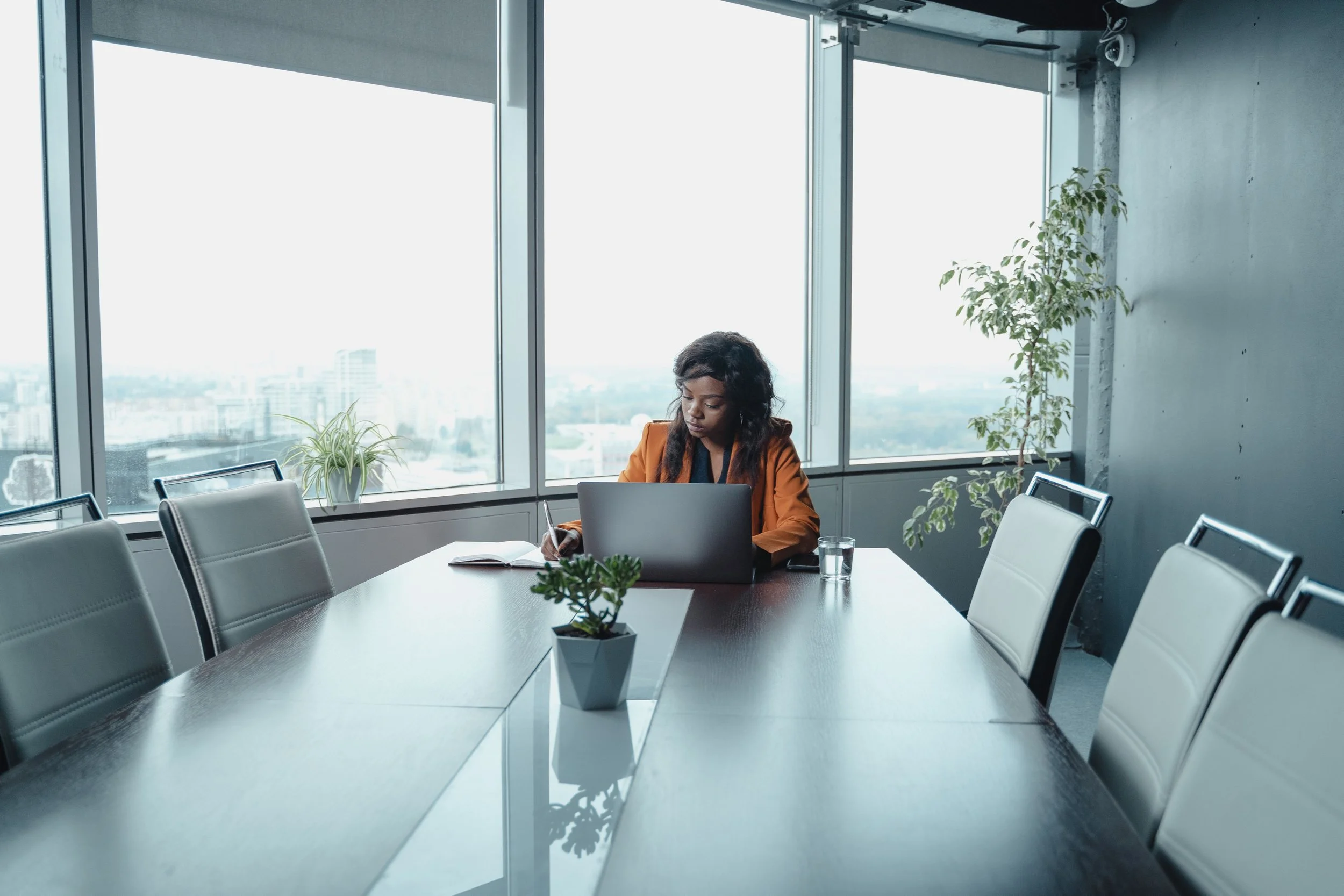 A woman with dark hair sitting at a conference table with a laptop, writing in a notebook, in a modern office with large windows and city view.