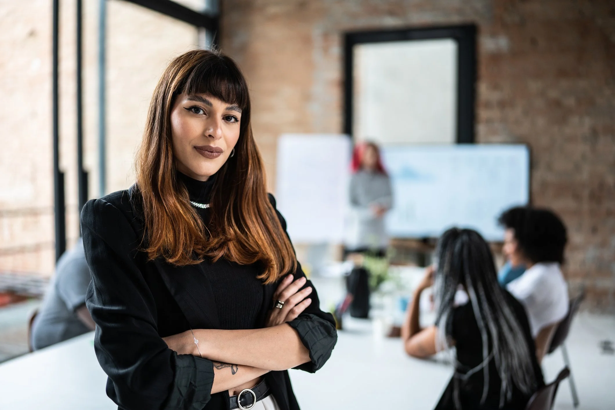 A woman with dark brown hair and bangs standing with her arms crossed in a modern office, with coworkers working at desks in the background.