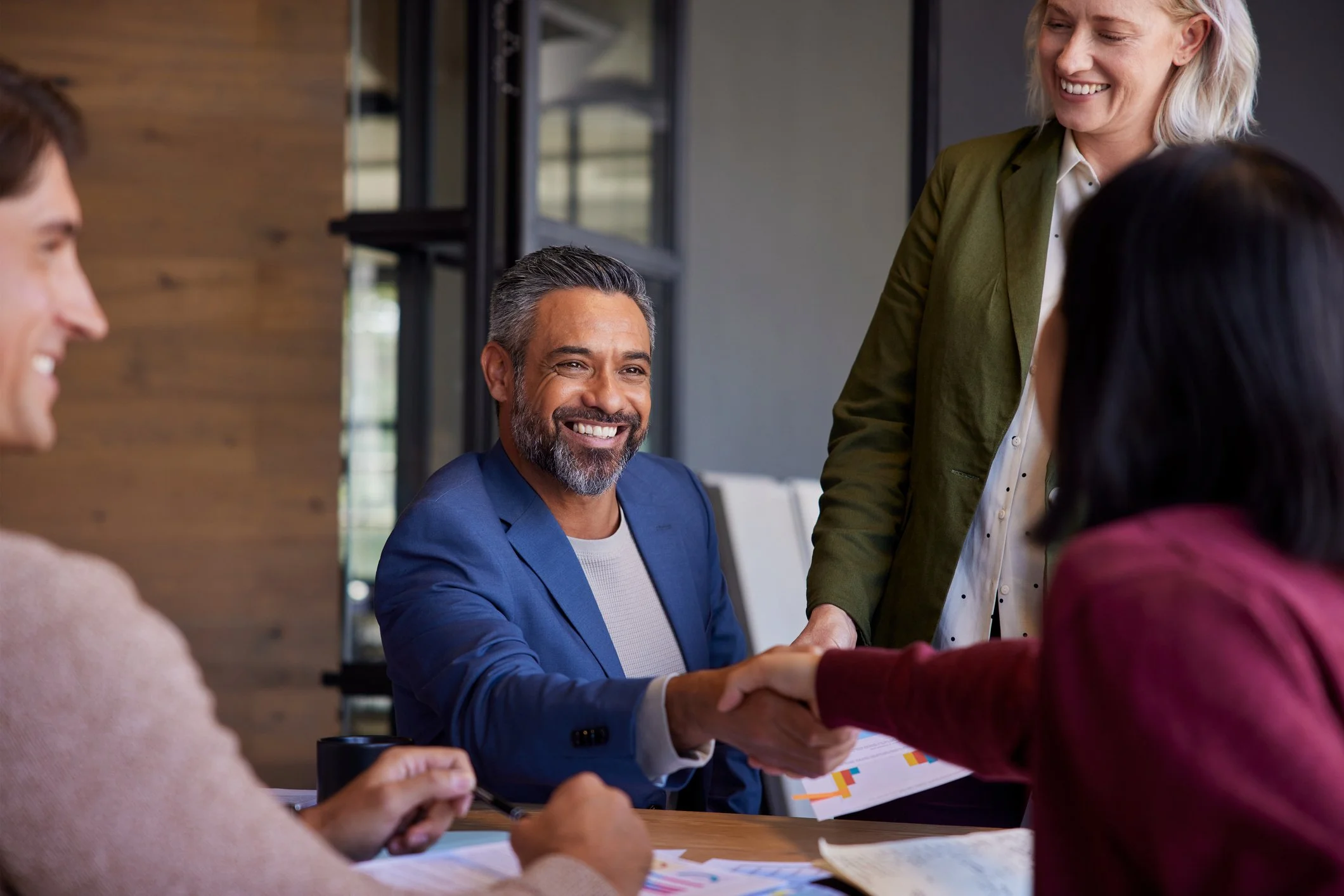 A professional meeting in a modern office, with diverse colleagues around a table. A man in a blue suit is shaking hands with a woman in a maroon top, smiling. Other coworkers are engaged, with documents and charts visible on the table.