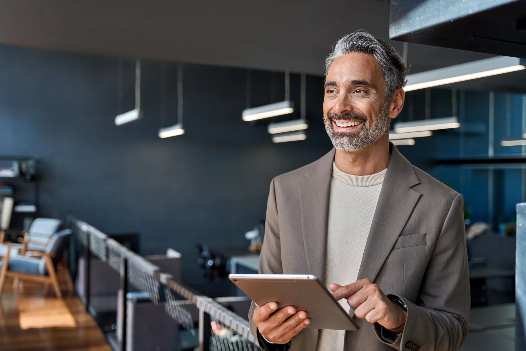 A smiling middle-aged man with gray hair and beard, wearing a beige suit jacket and white sweater, holding a tablet in an office or lounge area with modern furniture and hanging lights.