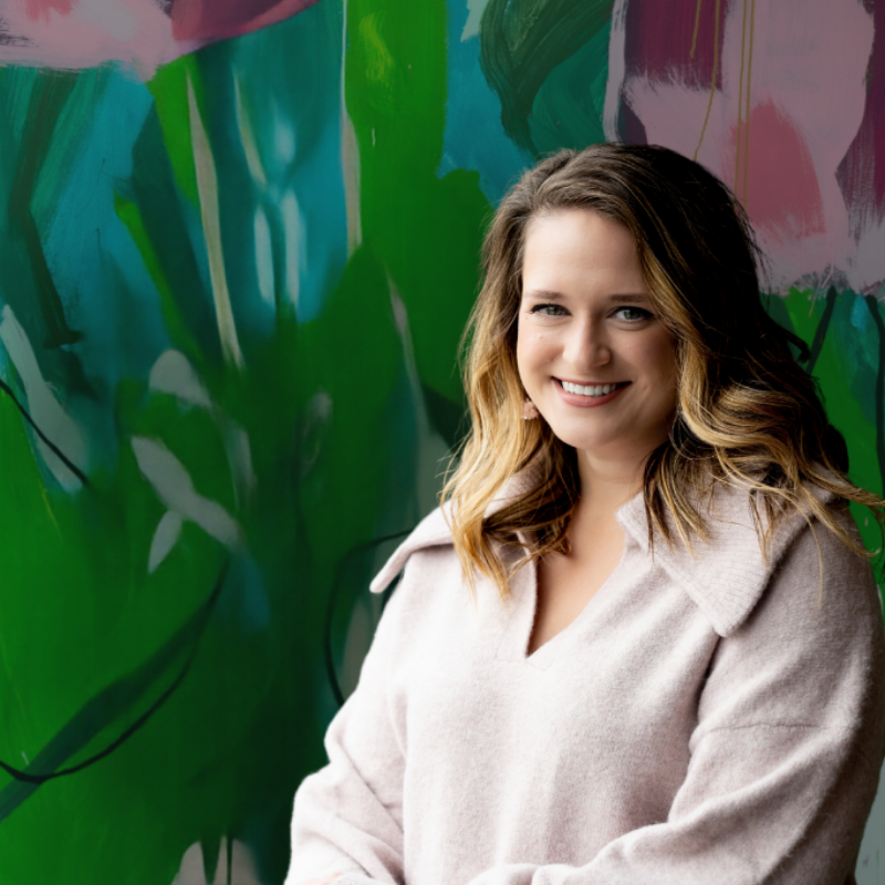 A woman with wavy blonde hair smiling and standing in front of a colorful abstract mural with green and pink hues.