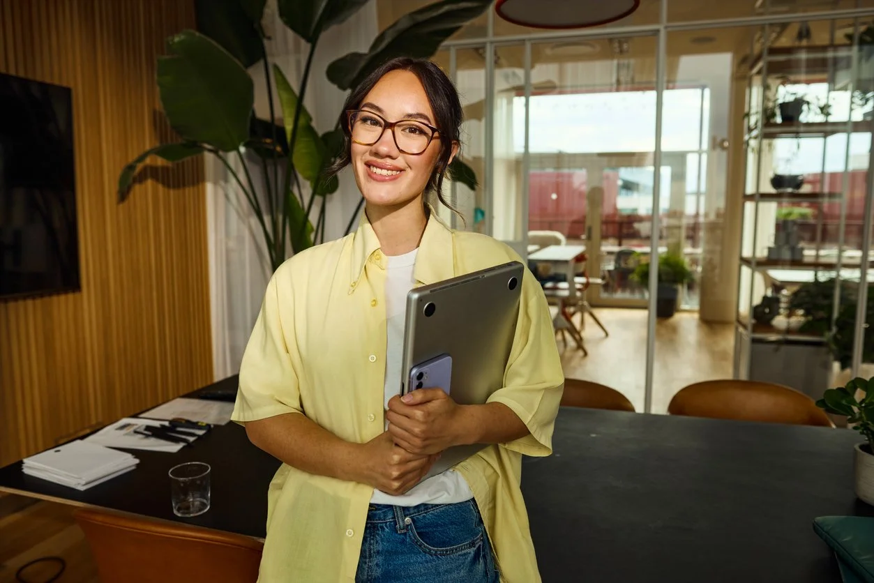 A woman wearing glasses and a yellow shirt, holding a laptop and smartphone, smiling in a modern office space with large windows, plants, and wooden accents.