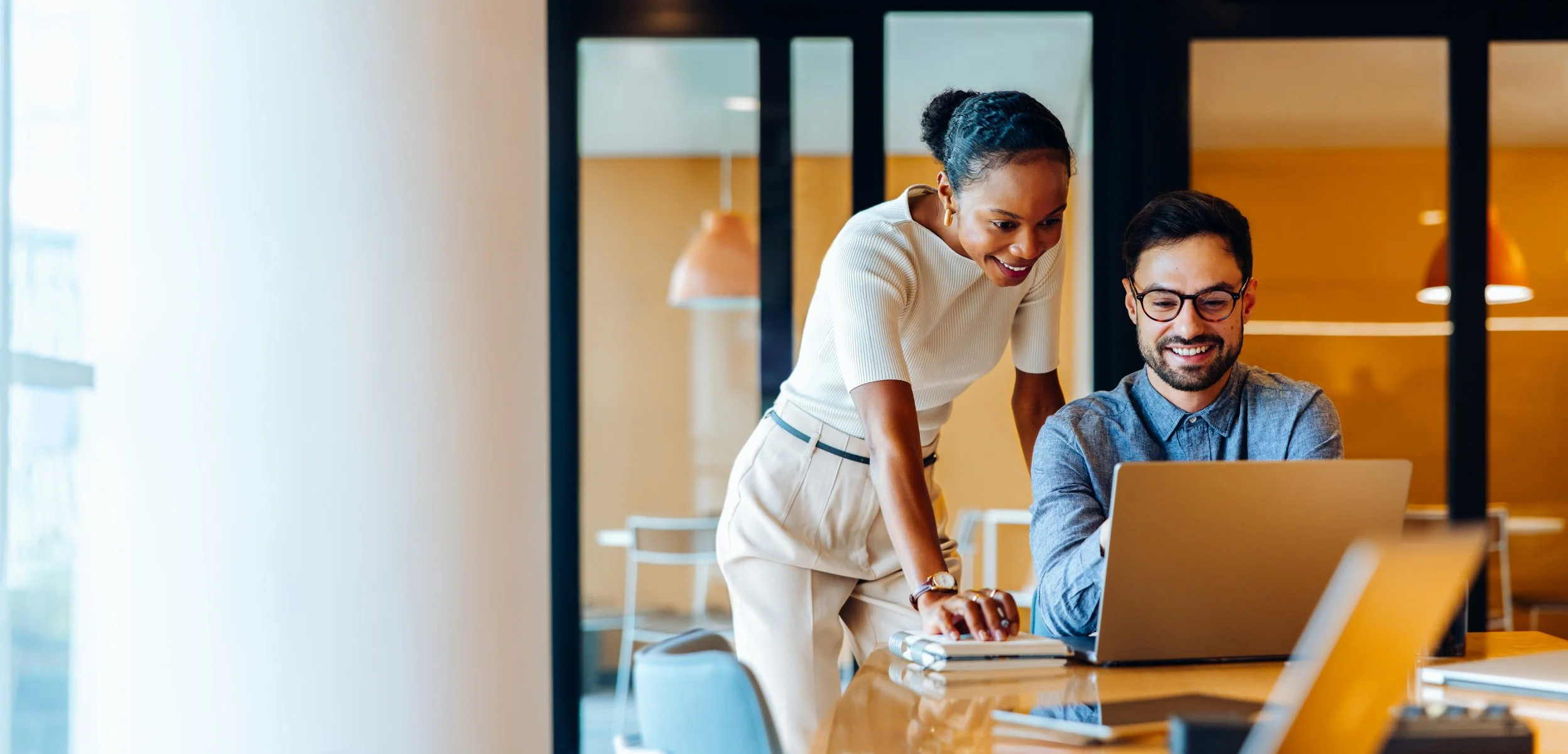 A woman and a man are working together at a laptop in a modern office, with the woman standing and leaning over the man's shoulder, both smiling.