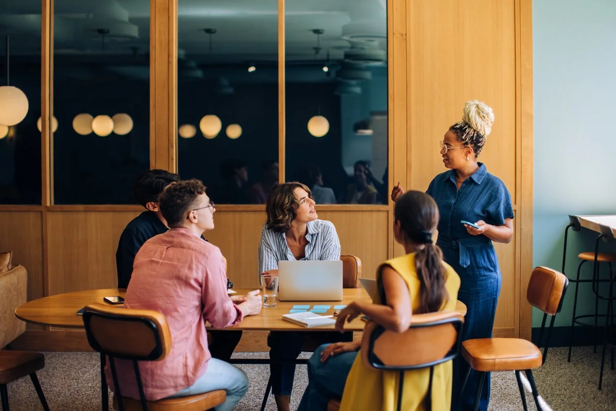 A group of five diverse people having a meeting in a conference room. One woman is standing and speaking to the others, who are sitting around a table with laptops, notebooks, and glasses of water.