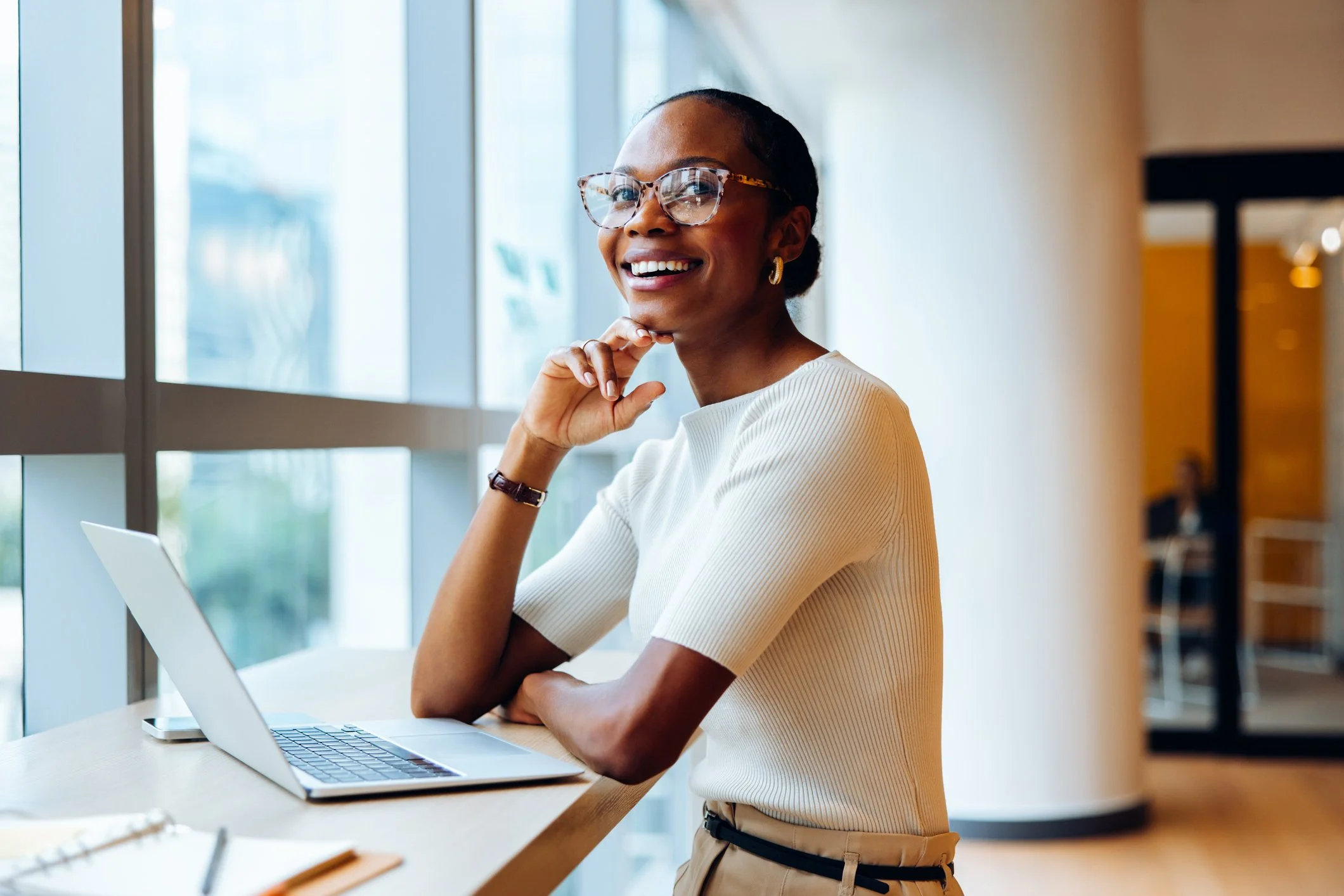 A smiling woman with glasses sitting at a table in front of a laptop in a bright indoor space with large windows.