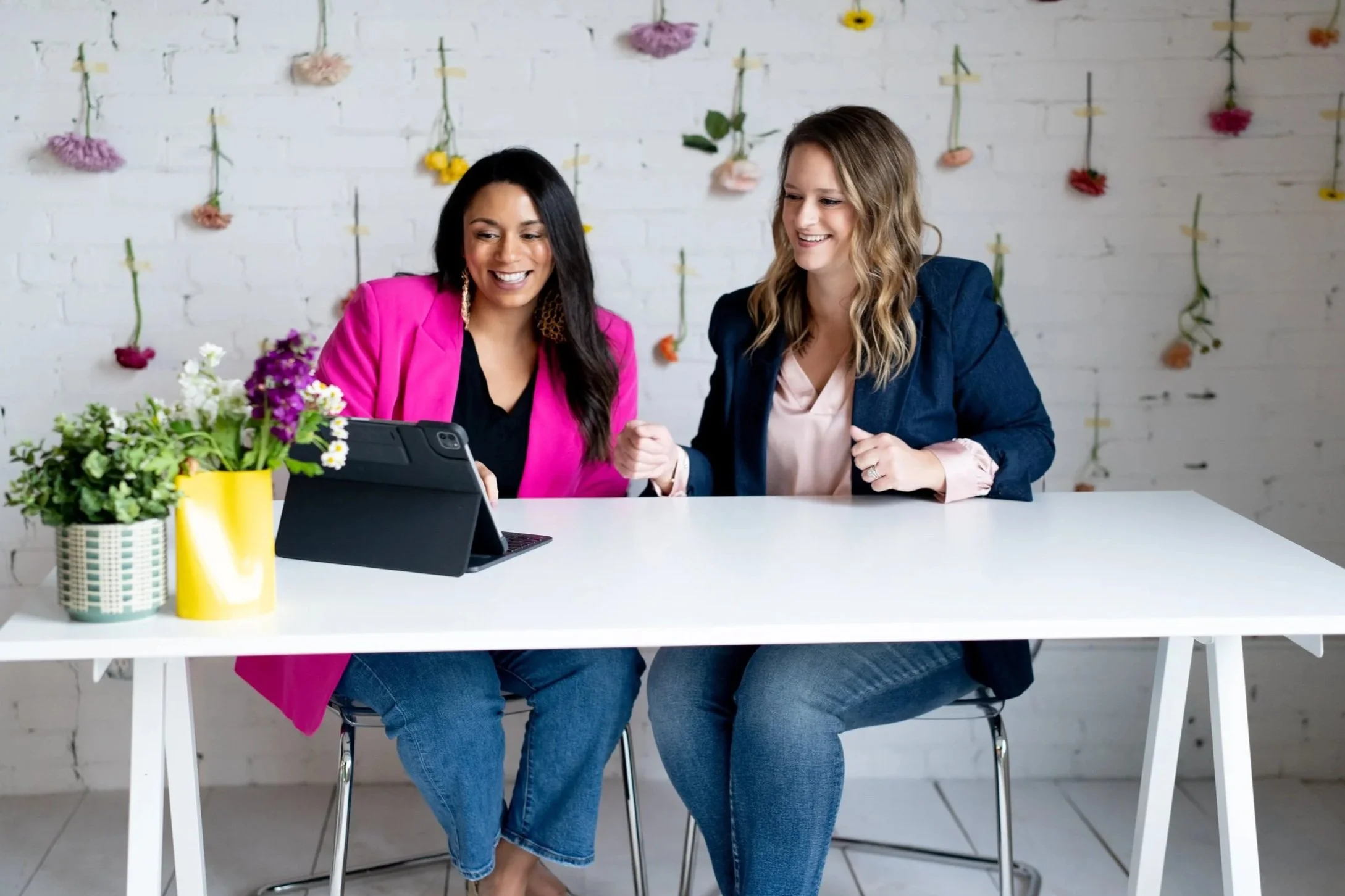 Two women sitting at a white table, looking at a tablet, with colorful flowers in front of them. The woman on the left has dark hair, wearing a pink blazer, and the woman on the right has blonde hair, wearing a dark blazer. There are flower decorations hanging on a white brick wall behind them.