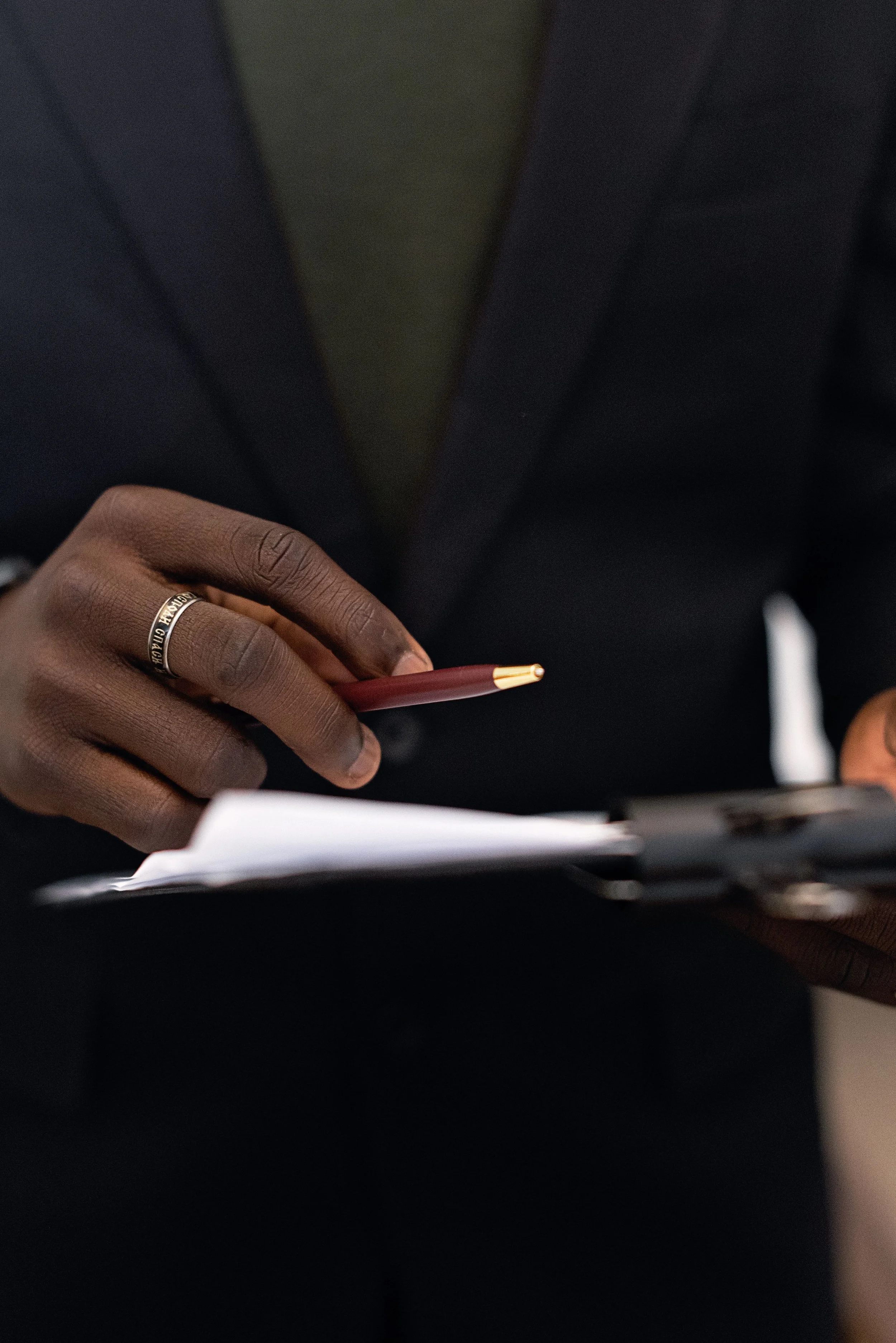 Close-up of a person's hand holding a pen, wearing a silver ring, with papers or a notebook in front, dressed in a dark suit.