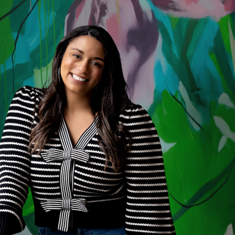 A young woman with long dark hair, smiling and wearing a black and white striped sweater with a bow detail on the front, standing in front of a colorful abstract mural.