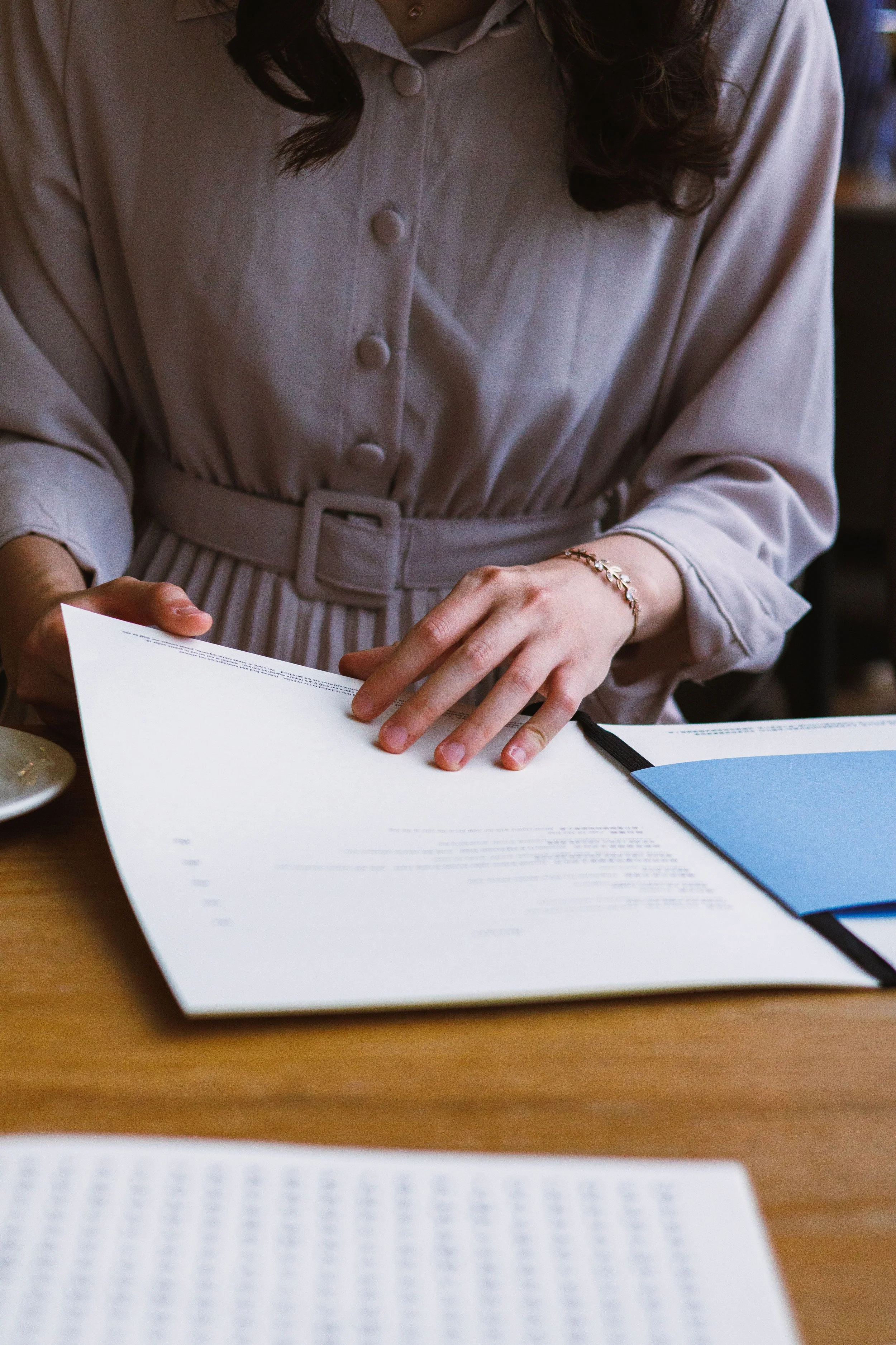 A woman dressed in a beige button-up shirt with a belt, sitting at a desk, holding and looking at a document or booklet with printed text. There is another document or booklet with blue cover on the desk.