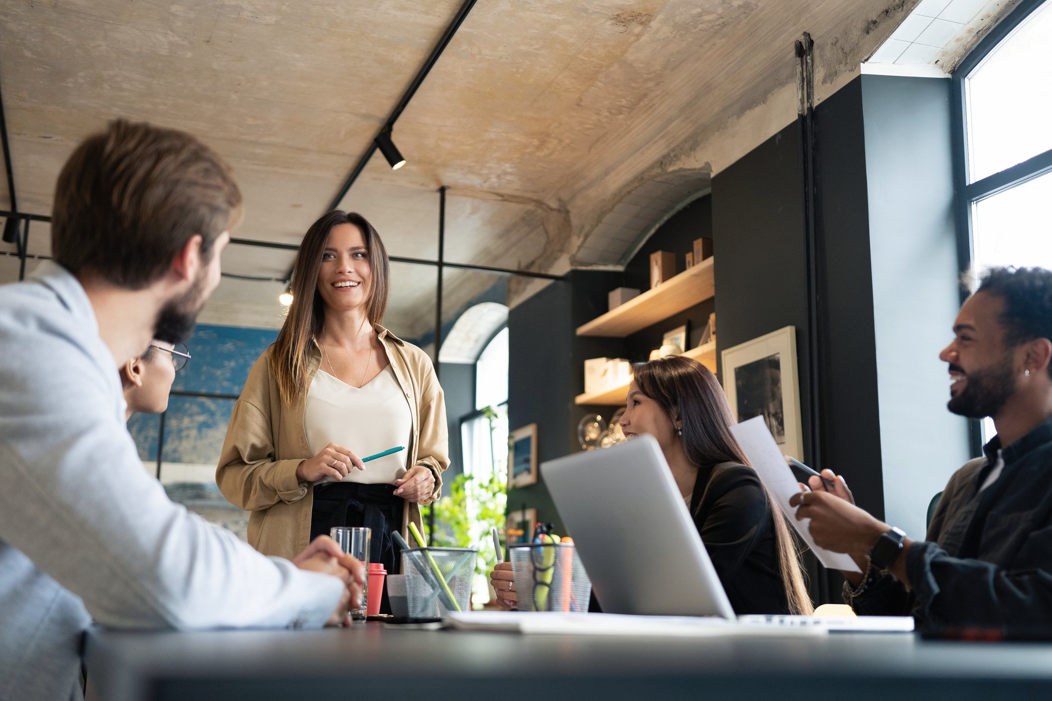 A diverse group of young professionals in a modern office, engaged in a discussion with a smiling woman standing while holding a pen, others seated around a table with laptops and papers, large windows with natural light in the background.