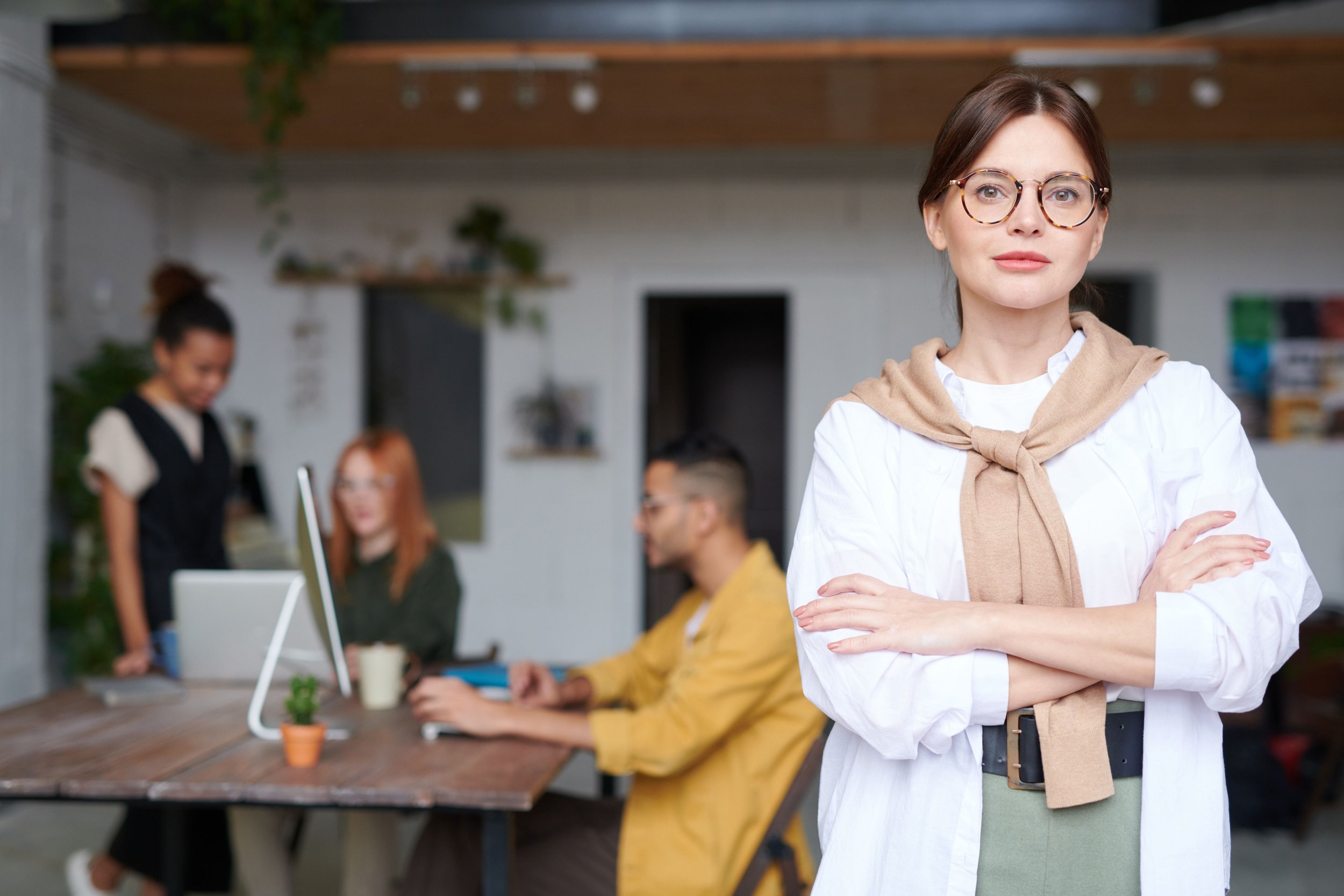 A young woman with glasses and a beige sweater tied around her neck standing with her arms crossed in a modern office space, with a group of people working at a table in the background.