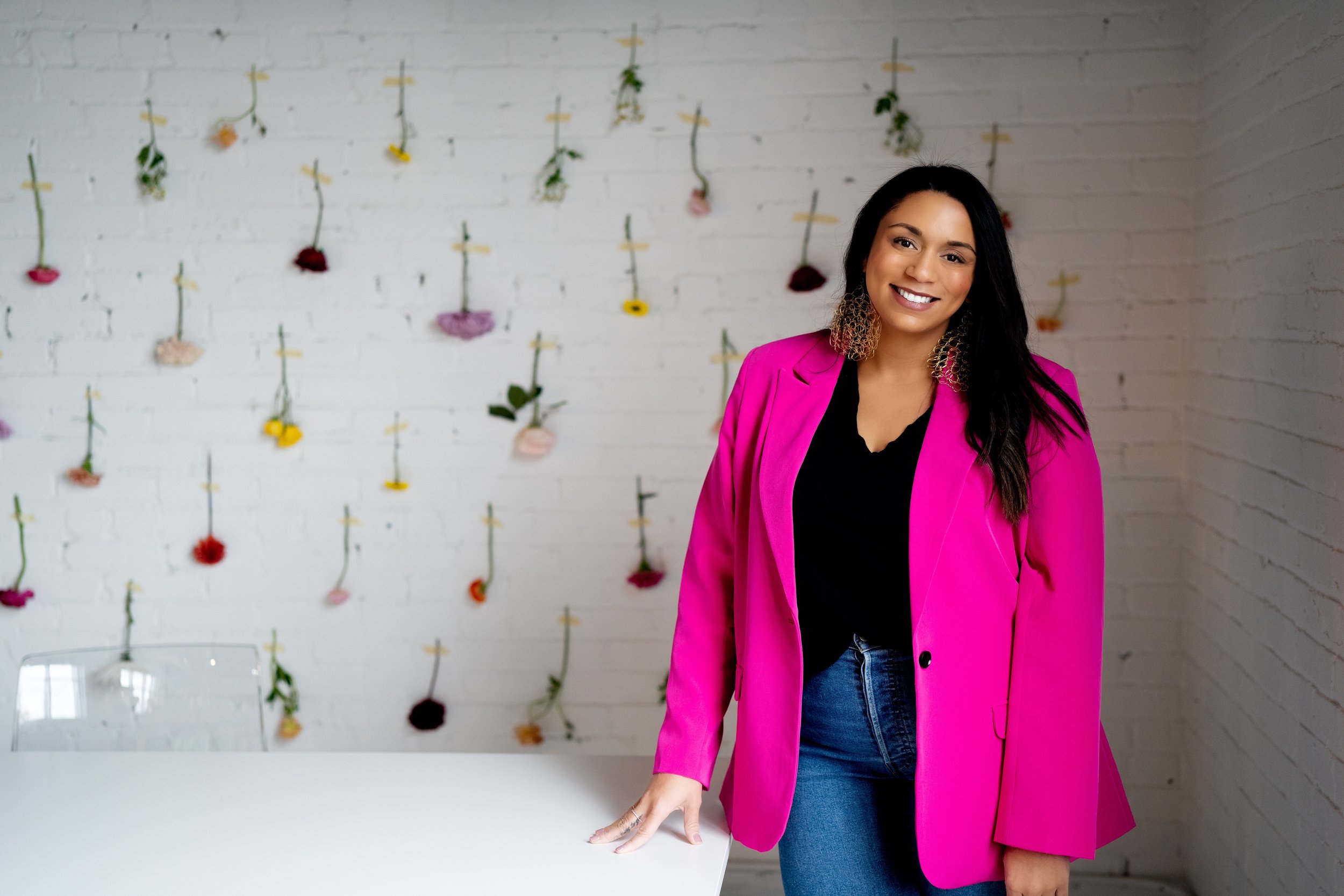A woman with long dark hair, wearing a bright pink blazer and dark top, smiling at the camera while standing in front of a white brick wall decorated with hanging flowers of different colors.