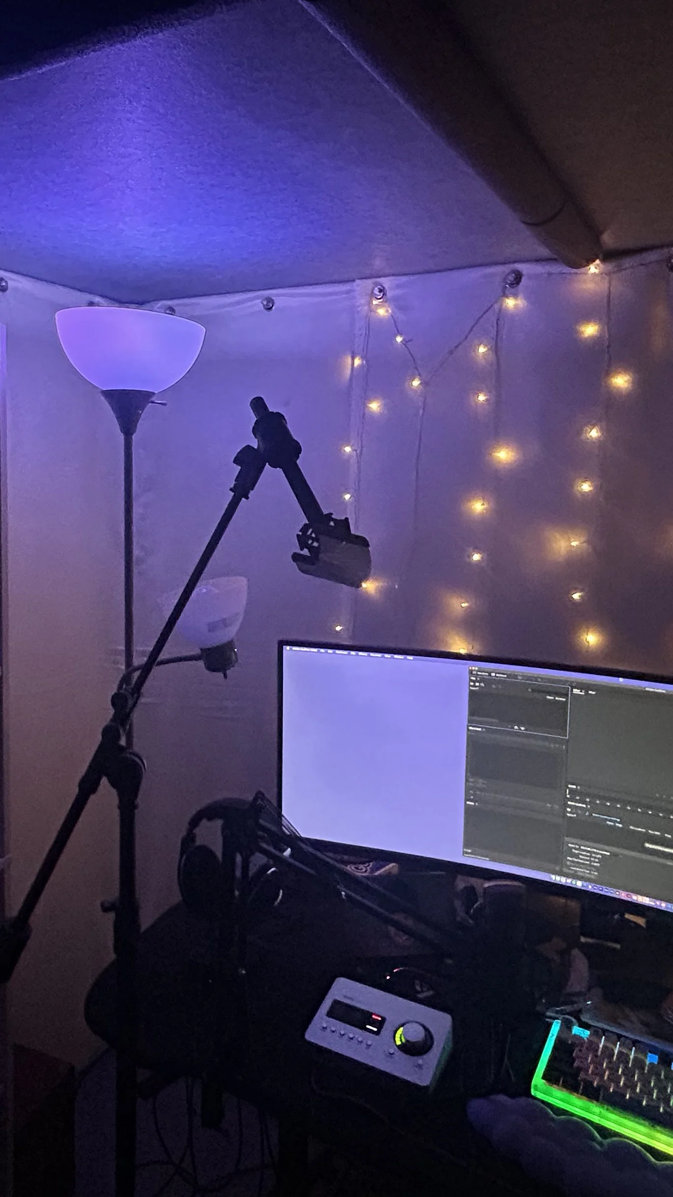 A workspace setup with a computer monitor displaying audio editing software, a microphone on a stand, headphones, and LED strip lights on the wall, with a tall floor lamp and fairy lights creating a cozy ambiance.