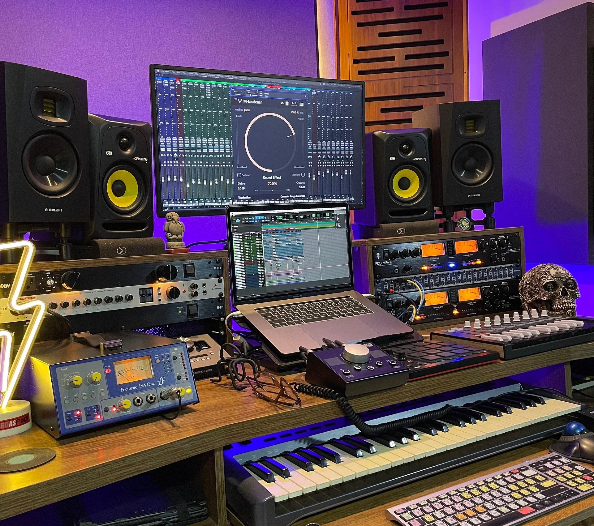 Music production studio workspace with studio monitors, mixing consoles, computer screens with digital audio workstation software, MIDI keyboard, synthesizers, audio interface, Skull decoration, and purple ambient lighting.