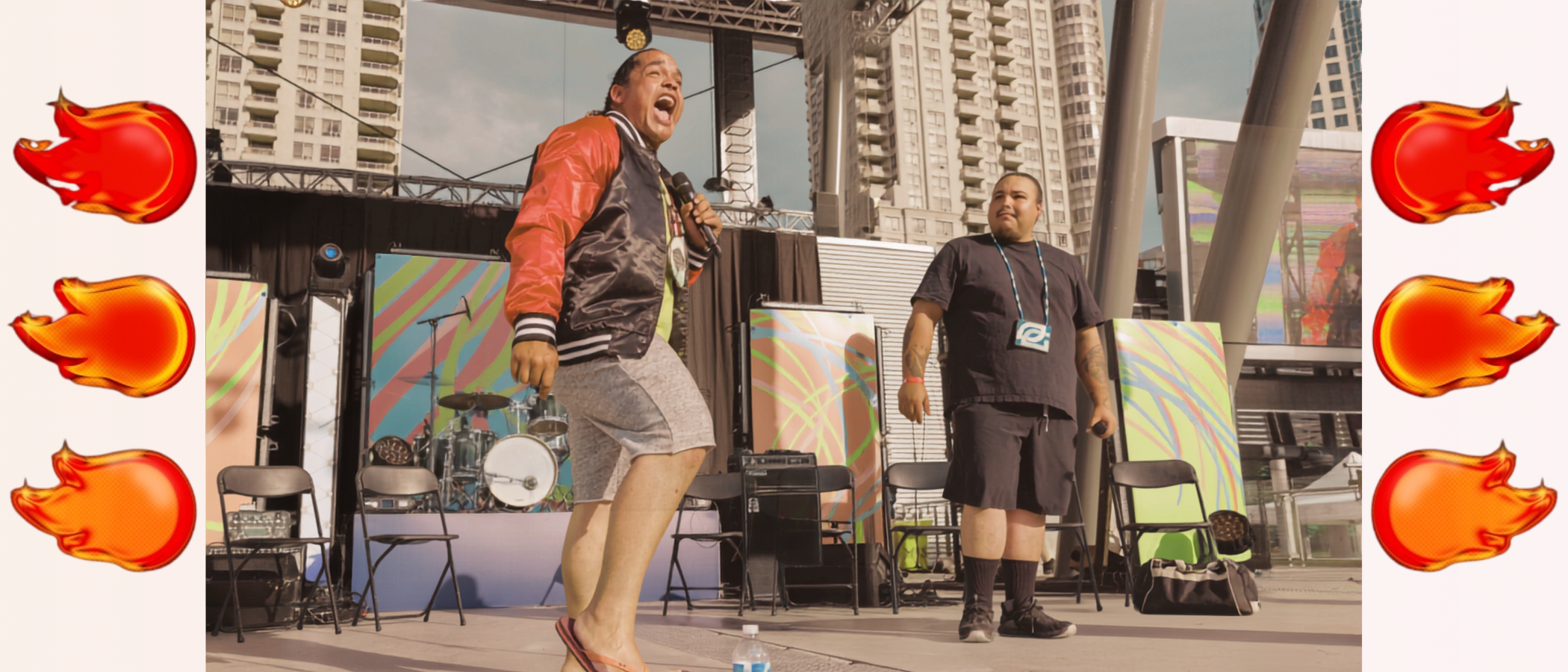 Two performers on an outdoor stage with a colorful background, surrounded by empty chairs and musical equipment, with tall buildings in the background.