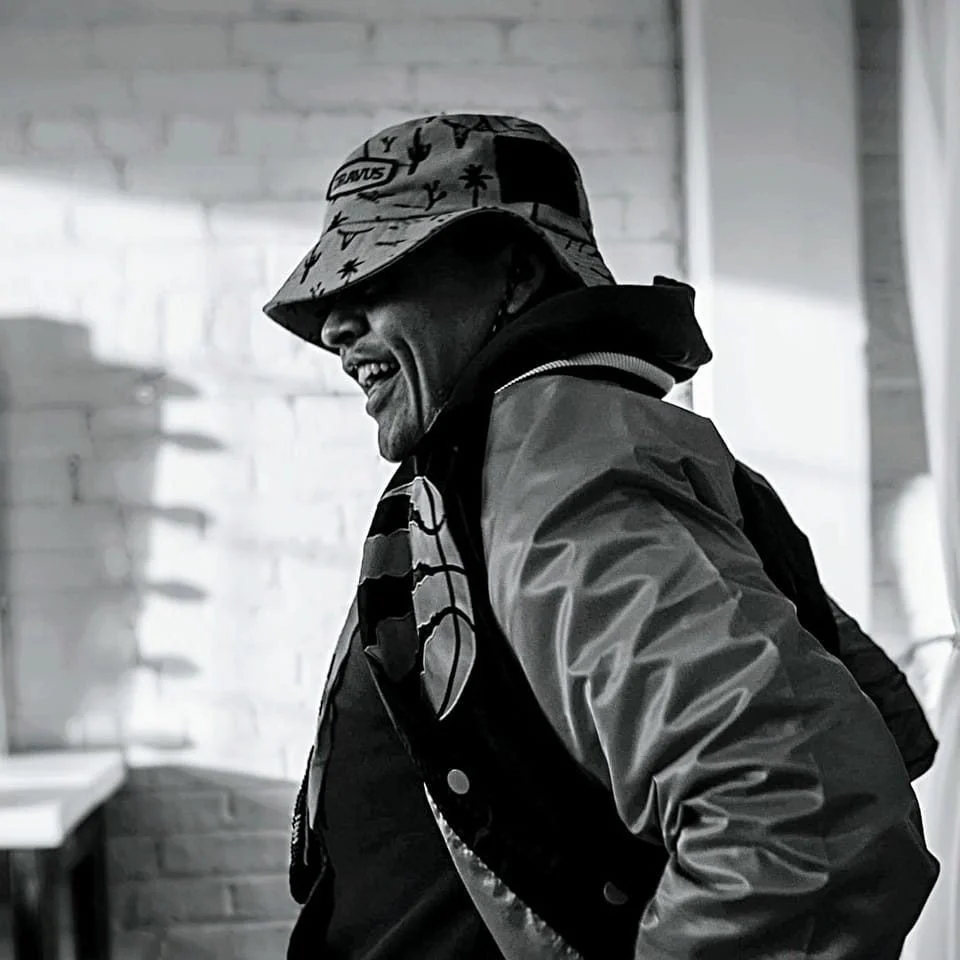 A man laughing, wearing a patterned bucket hat, a hoodie, and a jacket, standing in front of a white brick wall.