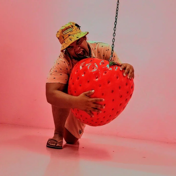Person wearing a bucket hat and orange shirt hugging a large, red, strawberry-shaped object hanging from a chain.