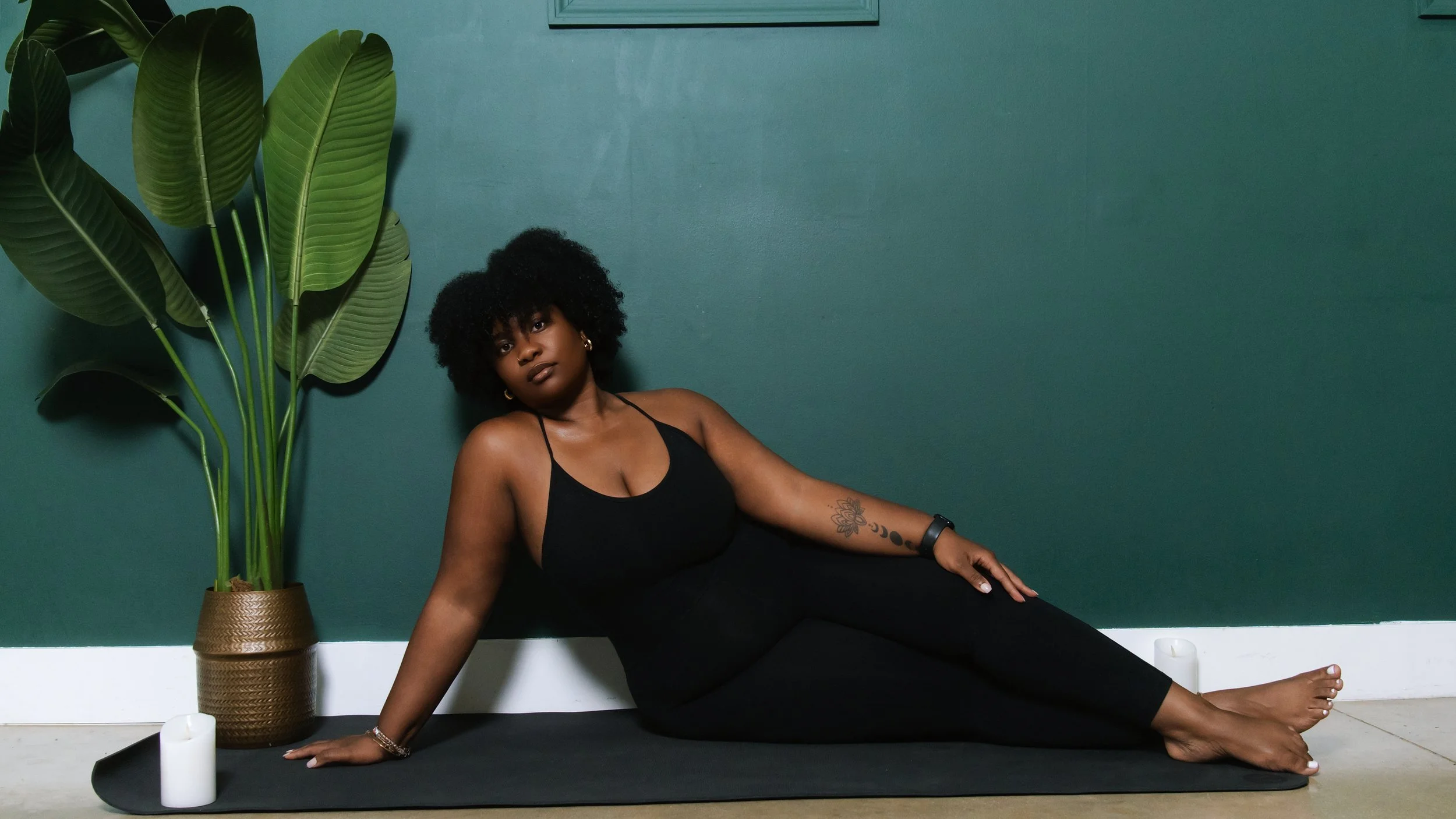 A woman with curly black hair and a tattoo on her arm doing yoga on a black mat in front of a teal wall, with large green plant leaves in a brown pot to her left.