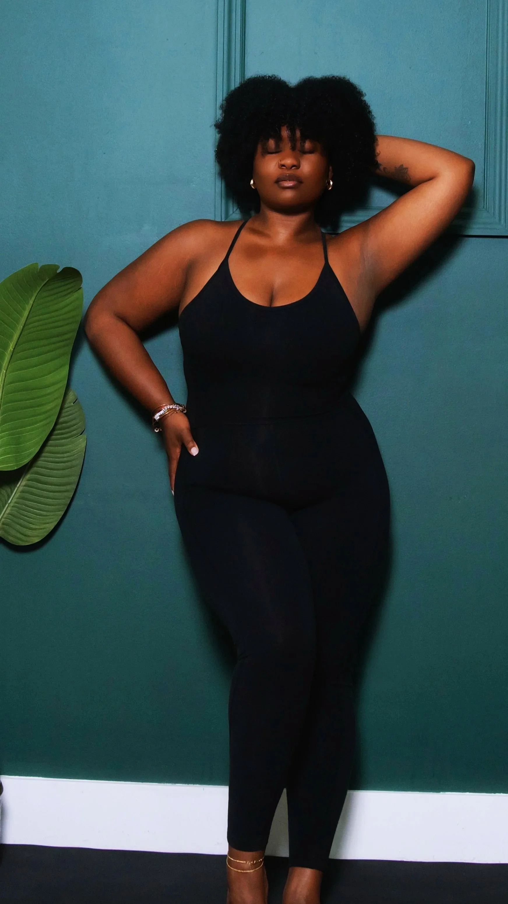 A woman with natural curly hair, wearing a black top and pants, stands against a dark green wall, with one arm behind her head and the other resting on her hip, next to a large green plant.