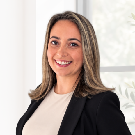 Portrait of a smiling woman in professional attire standing indoors near a window with greenery outside.