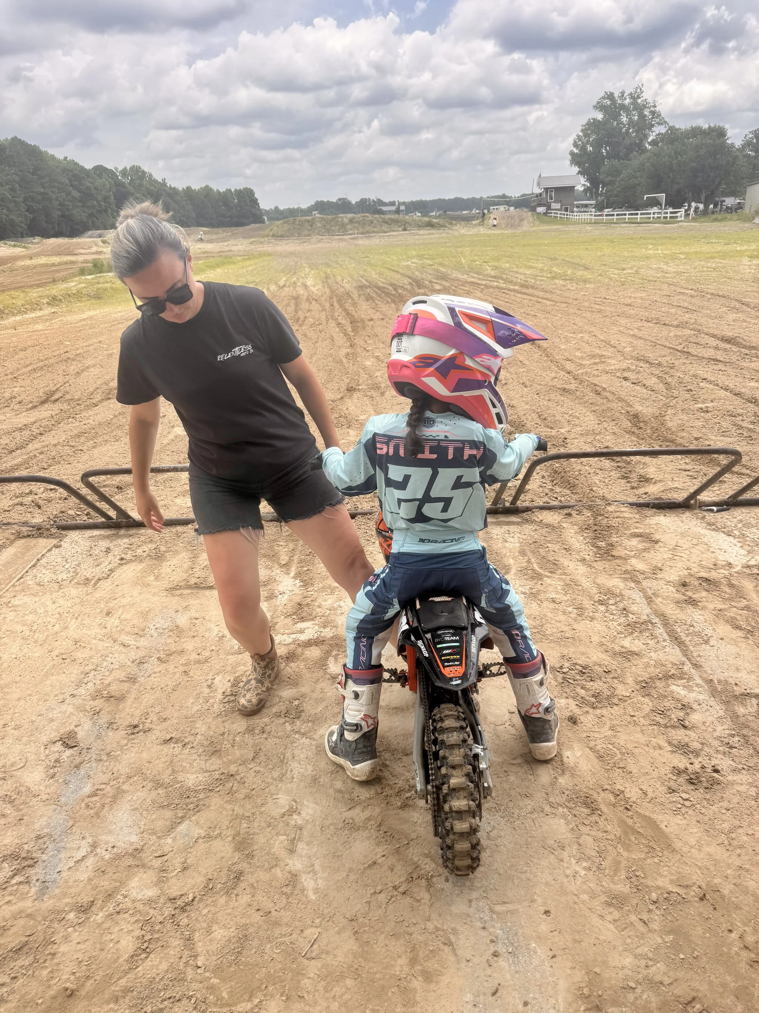 A young girl wearing a pink helmet sitting on a dirt bike with an adult woman standing beside her at an outdoor dirt track.