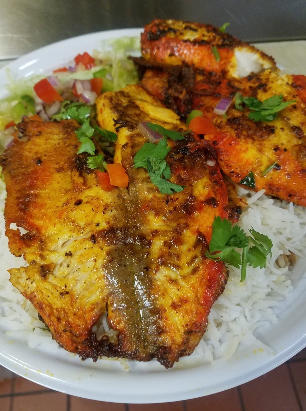 Close-up of a plate of white rice topped with grilled fish fillets, garnished with fresh cilantro, and accompanied by a small side salad with chopped lettuce, tomatoes, and onions.
