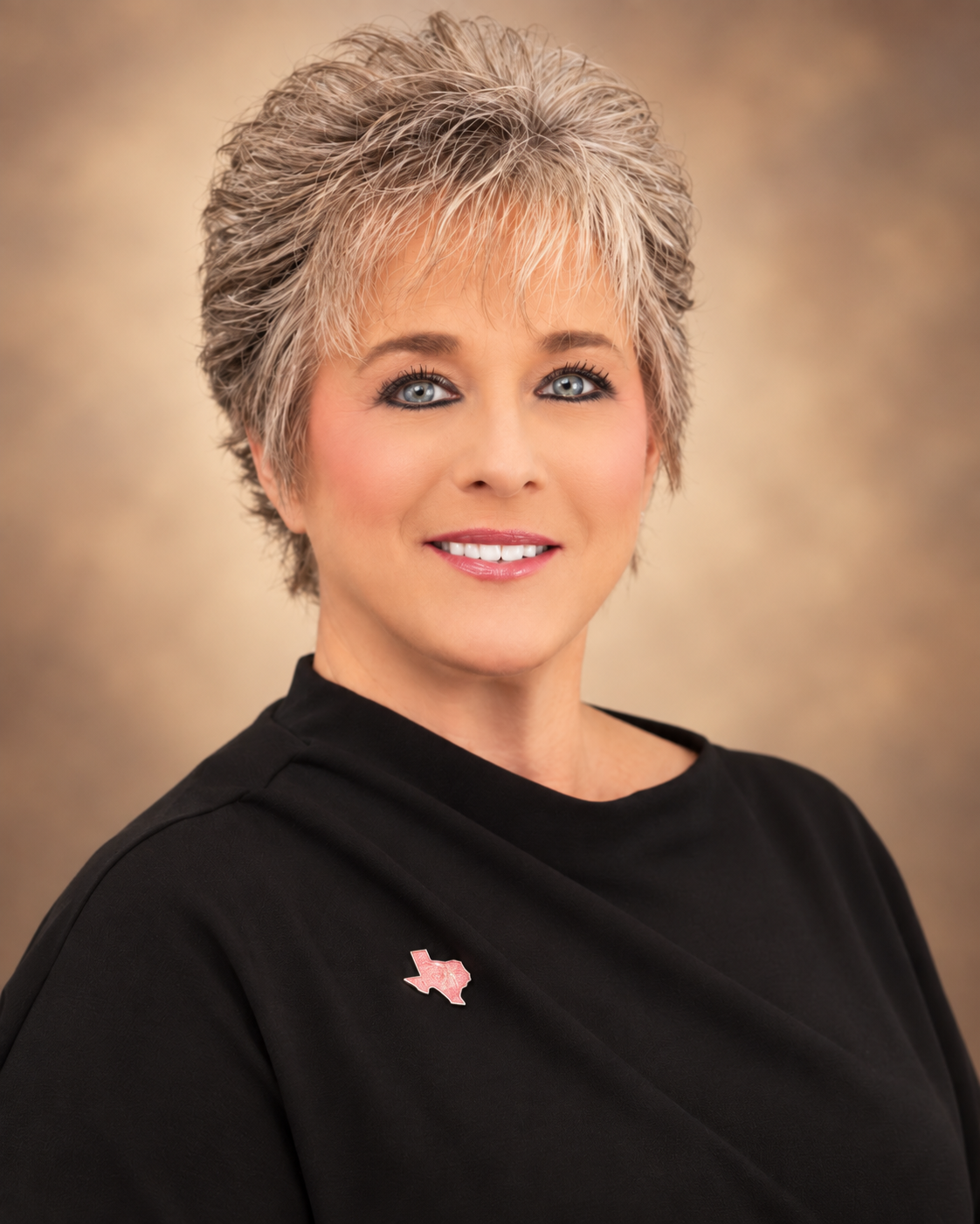 A professional woman with short, blonde, spiky hair and blue eyes, wearing a black top with a Texas-shaped pin on the shoulder, smiling against a beige background.