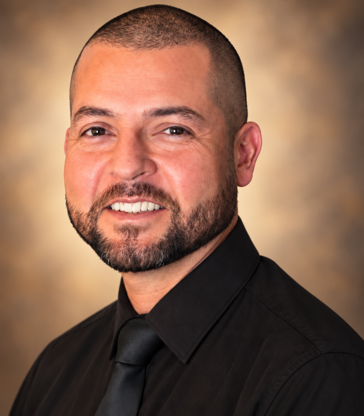 A man with short hair and a beard, dressed in a black shirt and black tie, smiling in a professional headshot.