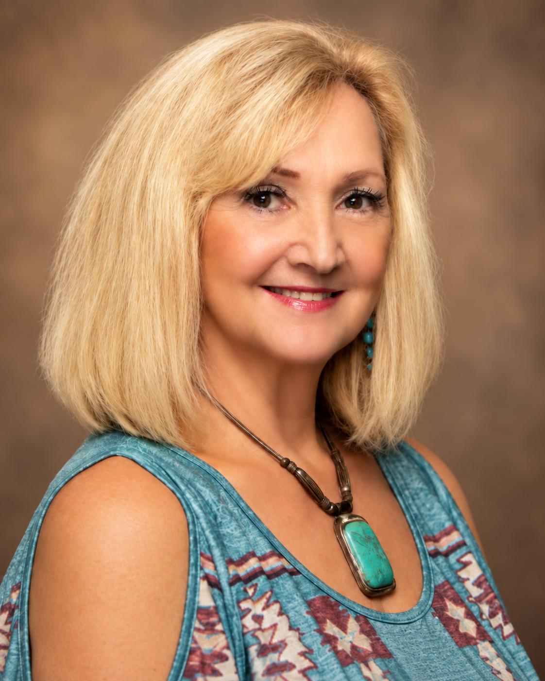 A middle-aged woman with blonde hair smiling at the camera, wearing a sleeveless turquoise top with tribal patterns, turquoise earrings, and a turquoise pendant necklace, against a neutral background.