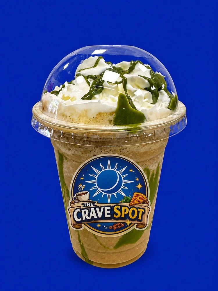 Iced coffee beverage topped with whipped cream, green syrup drizzle, and chocolate shavings in a clear plastic cup with the logo 'The Crave Spot' on a blue background.