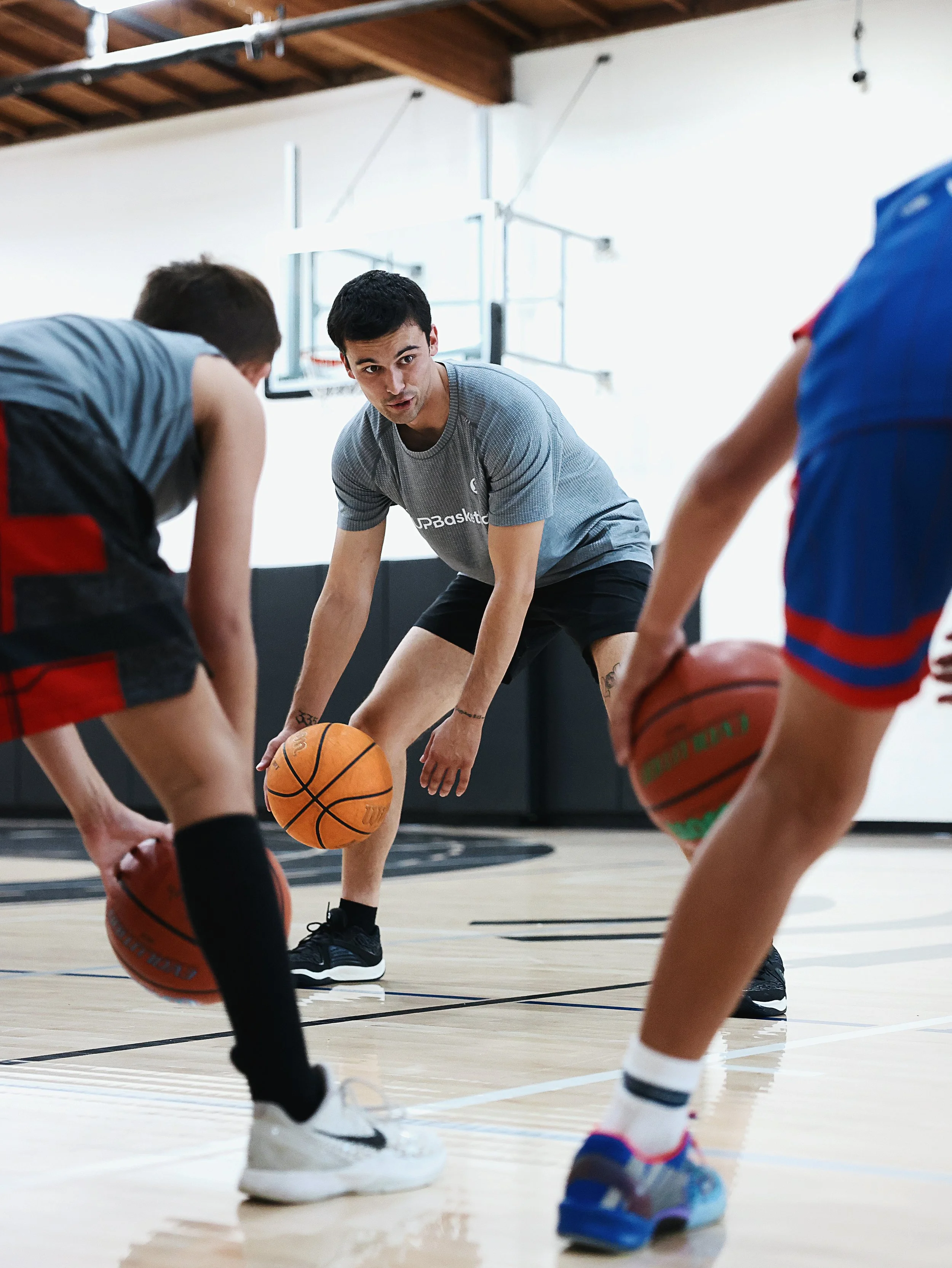 A basketball coach instructs young players during a practice in an indoor gym. The coach, dressed in a gray t-shirt and black shorts, is bending down with a basketball in hand, while the children, in sports gear, are gathered around him holding basketballs. The gym has a basketball hoop and a wooden ceiling.