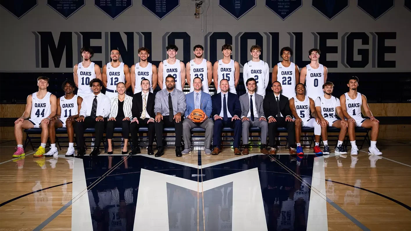 A basketball team and their coaches pose for a team photo in a gymnasium. The players are wearing white jerseys with 'OAKS' printed on them, and the coaches are dressed in formal attire. They are seated and standing in front of a large banner and a basketball court.