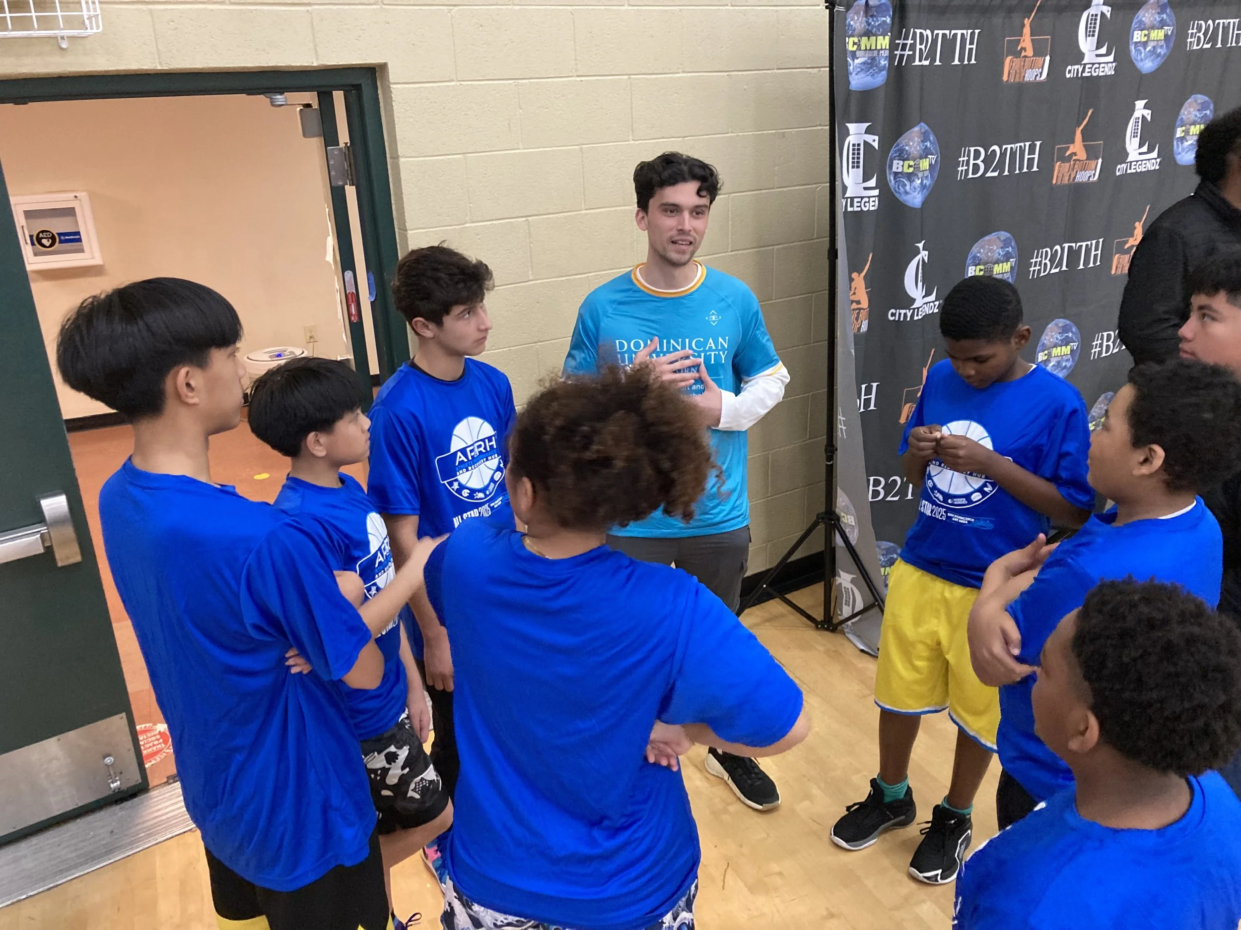 A group of young boys in blue sports shirts gathered around a coach who is speaking to them, indoors near a black backdrop with sponsorship logos and white text. The boys are listening attentively.