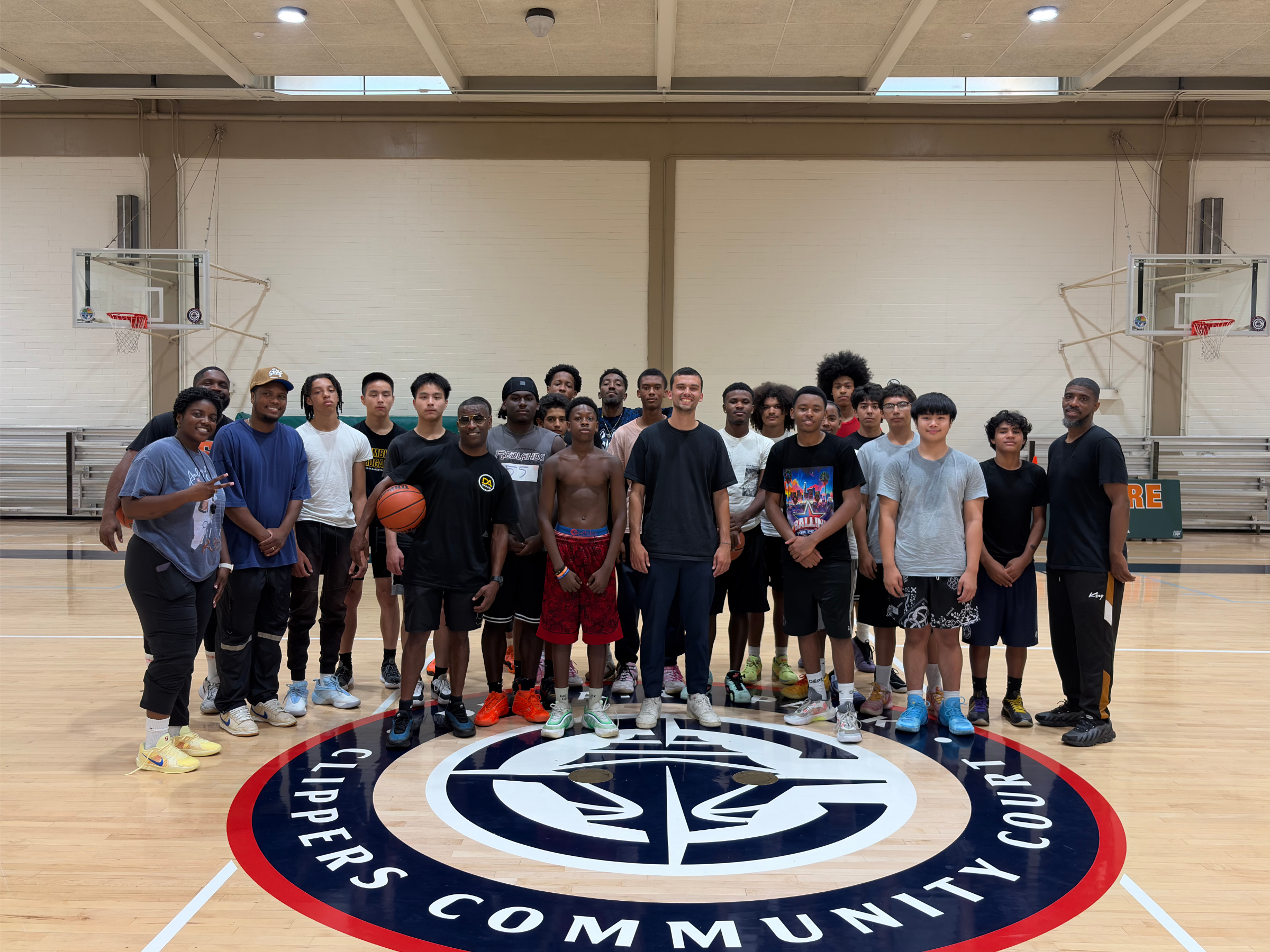 Group of diverse young men and women posing together on an indoor basketball court, some holding basketballs. They are standing around a large circular logo on the floor that says 'City of Commerce'.